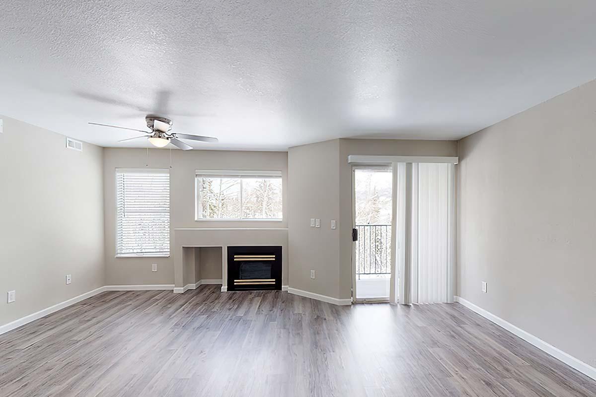 Spacious, empty living room with light-colored walls and wood-like flooring. Features a ceiling fan, large windows allowing natural light, a cozy fireplace in the corner, and a sliding glass door leading to a balcony. The room is designed for comfortable living and abundance of space.