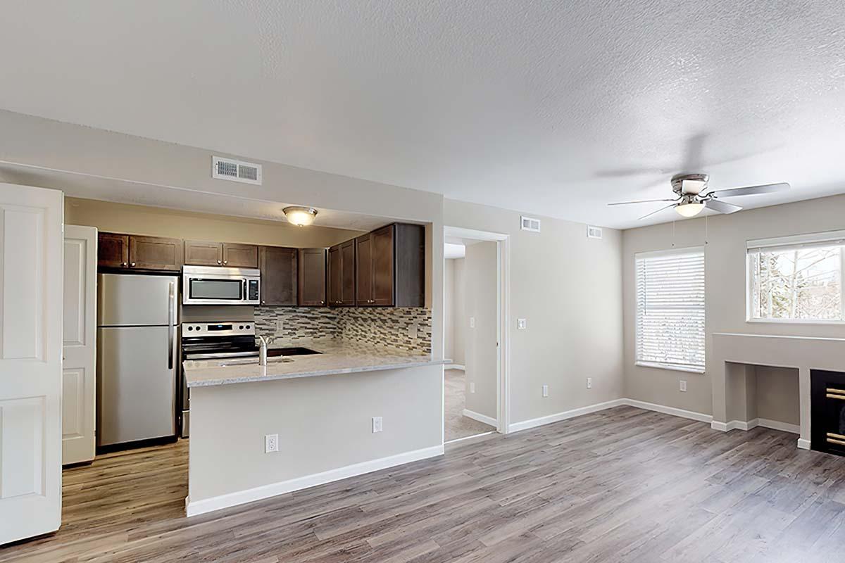 Spacious living area featuring a kitchen with modern appliances, including a refrigerator and microwave. The room has light-colored walls, wood-like flooring, a ceiling fan, and large windows providing natural light. A small fireplace is visible in the corner, adding warmth to the space.