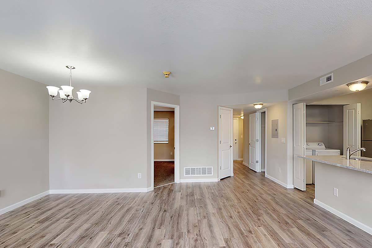 A spacious, modern living area with light brown wooden flooring. A doorway leads to another room, and there is a light fixture with three bulbs hanging from the ceiling. The walls are painted in a soft neutral color, and doorways can be seen on either side of the space, providing access to other areas of the apartment.