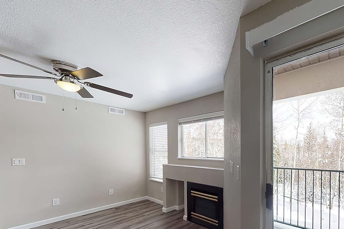A cozy living room featuring a ceiling fan, large window with blinds, and a faux fireplace. The walls are painted a neutral color, and the flooring is light wood. A balcony door leads to an outdoor space, and there is ample natural light illuminating the area.