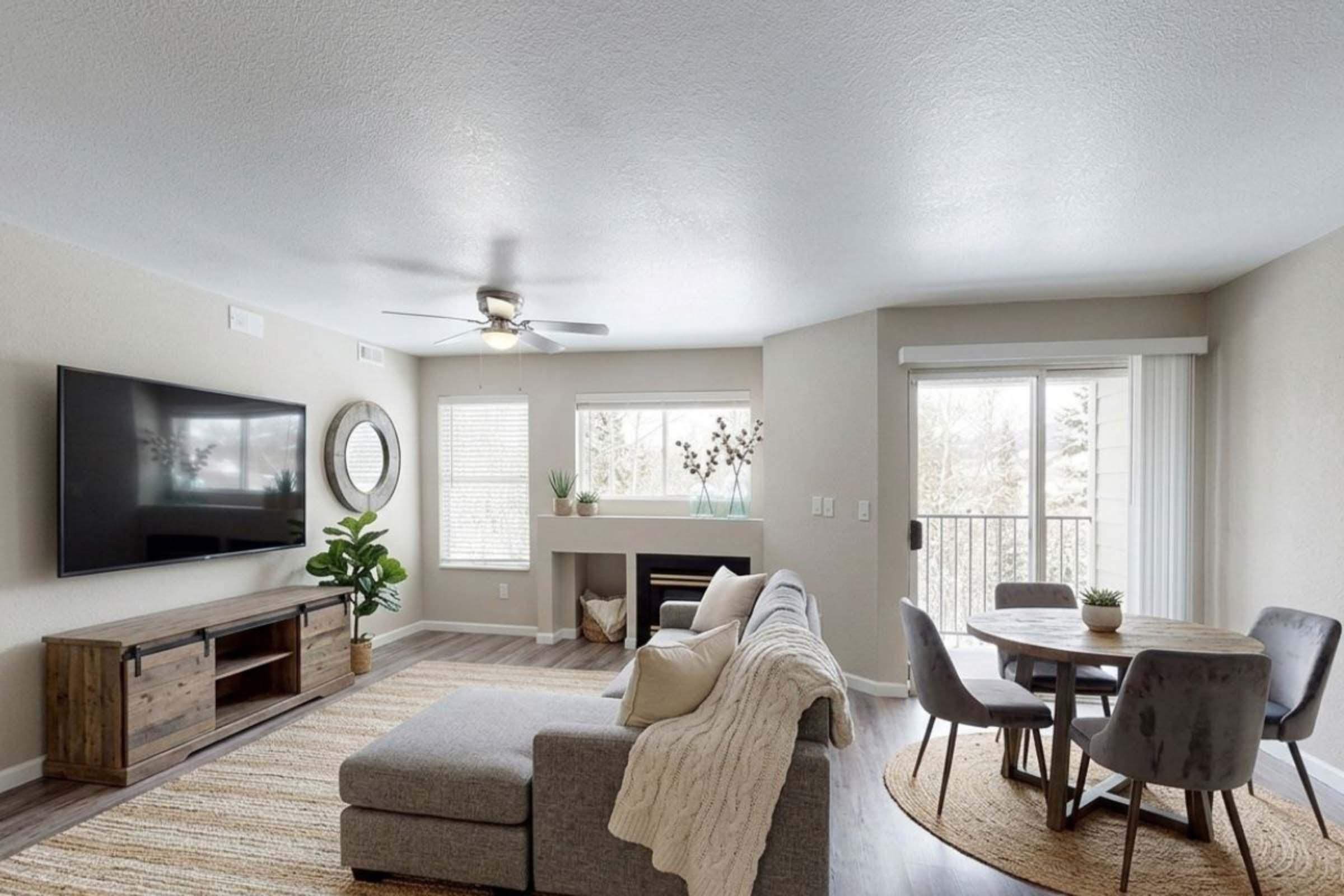 Modern living room featuring a comfortable gray sofa with a cozy throw, a wooden TV stand with a large flat-screen TV, and a ceiling fan. A circular wooden dining table with gray chairs sits nearby. Large windows provide natural light, and the space includes decorative plants and a neutral color palette for a warm ambiance.
