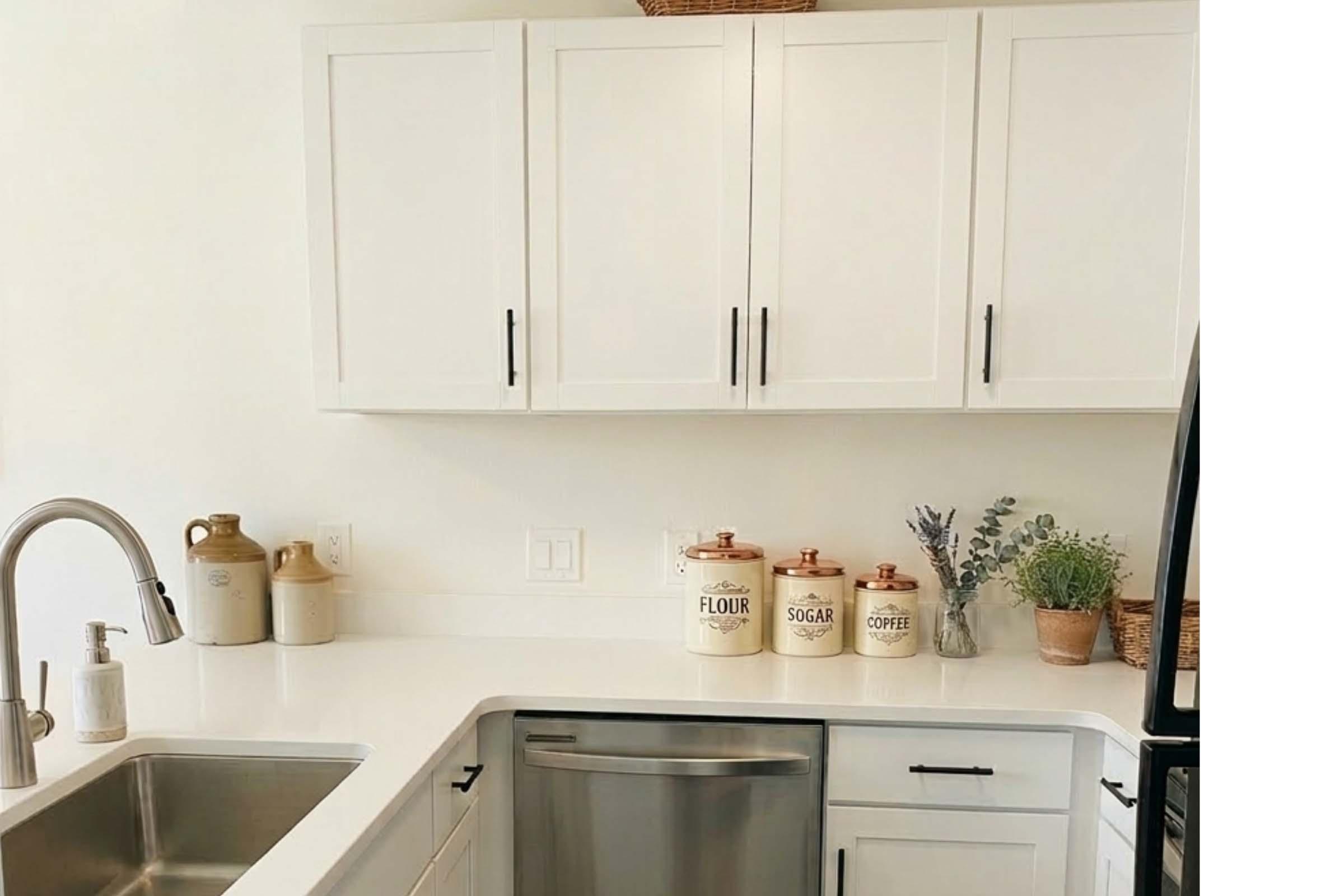 A modern kitchen with white cabinets and a stainless steel sink. The countertop features decorative jars labeled "FLOUR," "SUGAR," and "COFFEE," along with a small potted plant and a basket. The overall design is clean and minimalistic, emphasizing a bright and airy atmosphere.