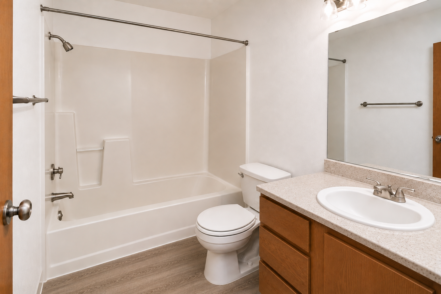 A clean, modern bathroom featuring a bathtub with a shower, a toilet, and a sink with a granite countertop. The walls are a light color, and there is a mirror above the sink. The floor has a wood-like appearance, and there are wooden cabinets beneath the sink.