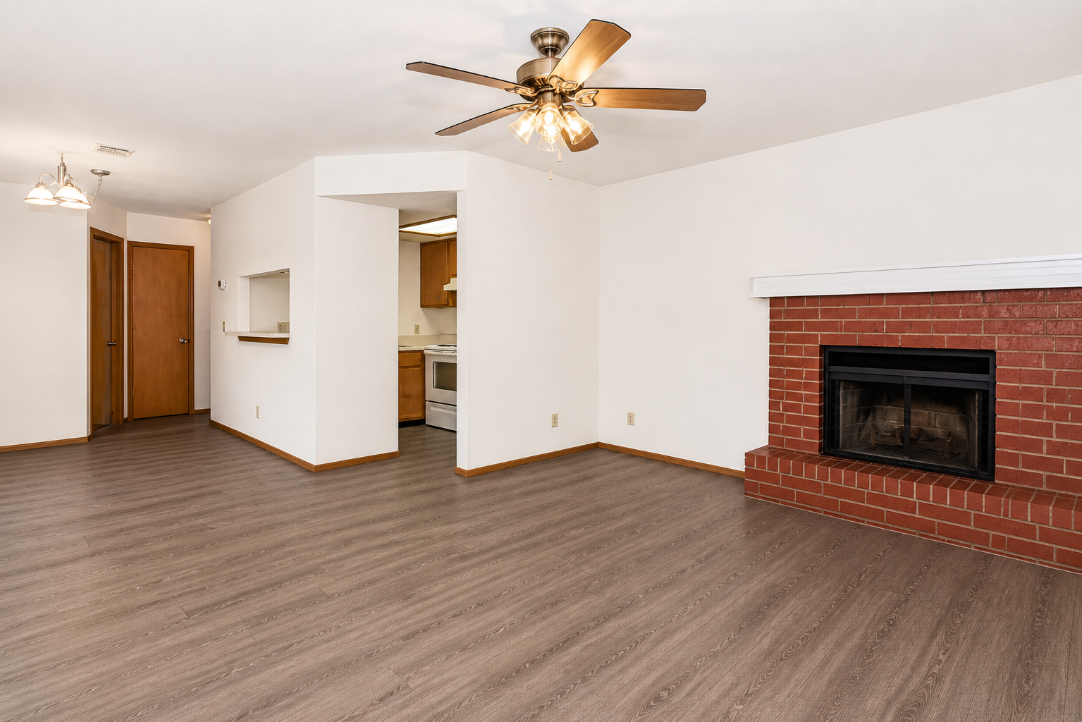 A spacious living room featuring hardwood-style flooring, a ceiling fan, a brick fireplace, and a doorway leading to a kitchen. Soft, neutral-colored walls and light fixtures add to the bright atmosphere of the space. The layout allows for easy movement and arrangement of furniture.