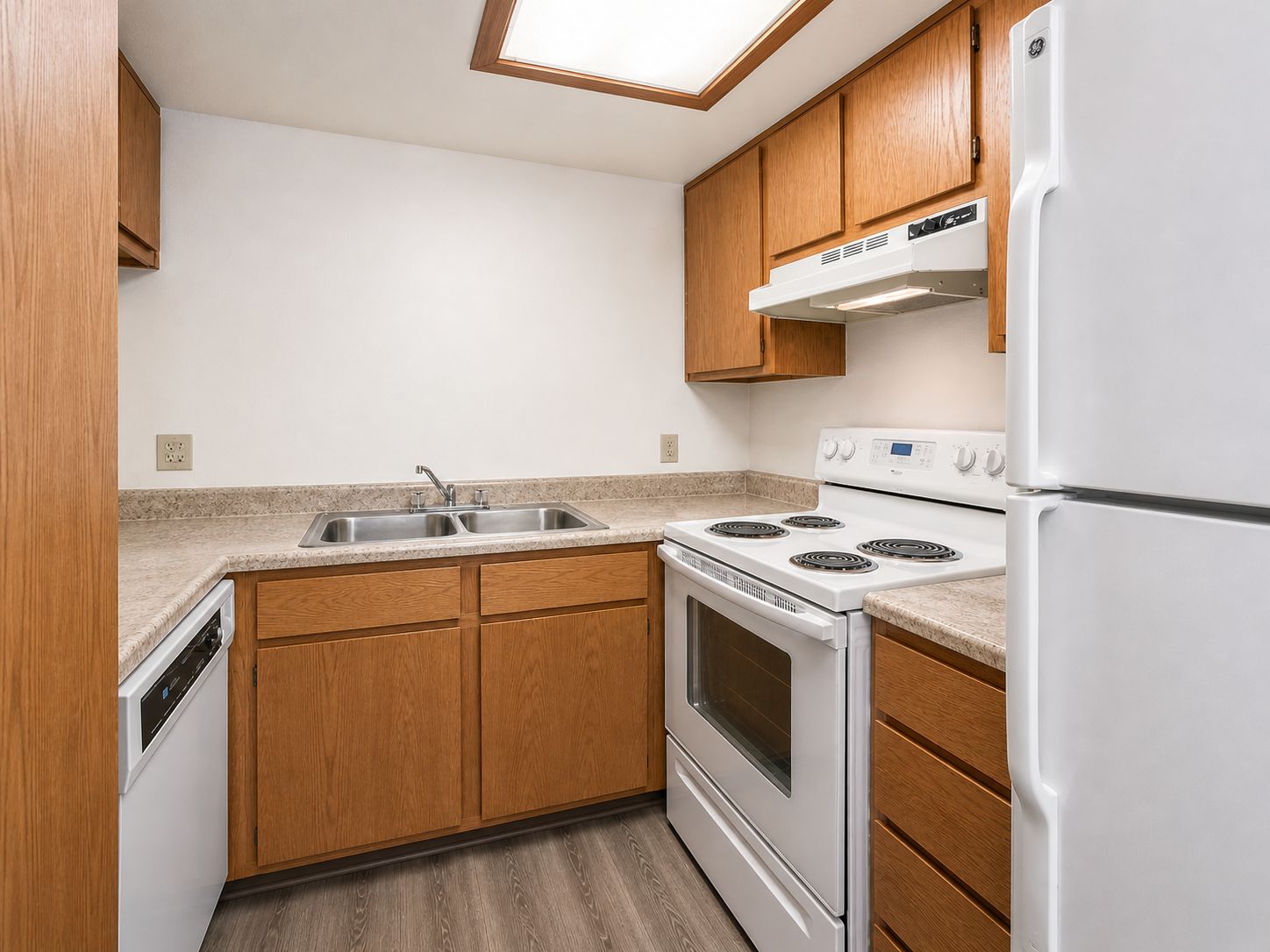 A small kitchen featuring wooden cabinets, a double sink, a white stove and oven, a microwave above, and a refrigerator. The countertop is light-colored, and there are four burners on the stove. The flooring is a light wood laminate, creating a warm and functional cooking space.