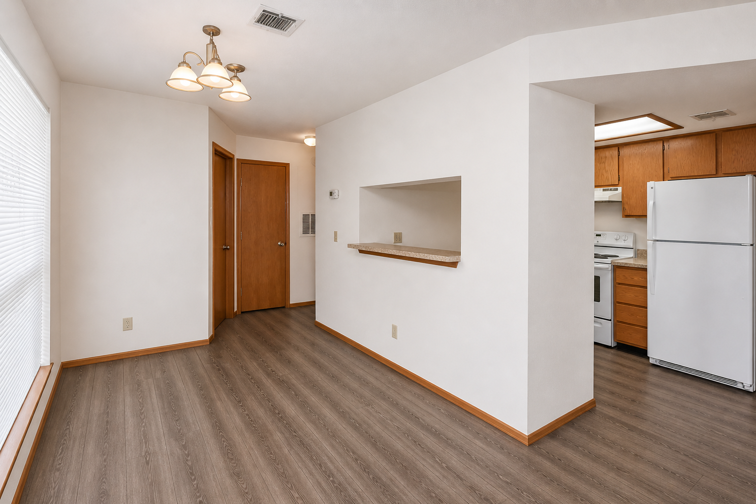 A modern, spacious interior featuring a living area with wood-like flooring, a small partition leading to a kitchen with wooden cabinets and white appliances. Natural light enters through a large window, and there are light fixtures hanging from the ceiling. The walls are painted a neutral color, creating a bright, open feel.