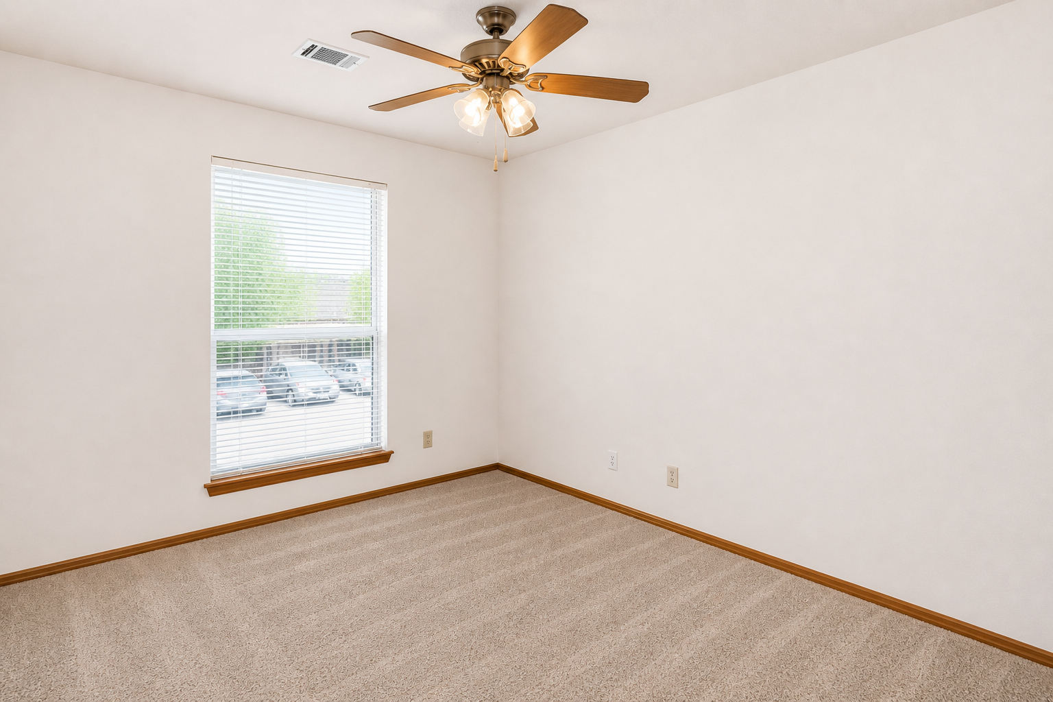Empty room with light-colored walls and a ceiling fan. The floor is carpeted in beige, and there's a window with blinds letting in natural light. A view of parked cars is visible outside the window, and the space appears clean and fresh, suitable for various uses.