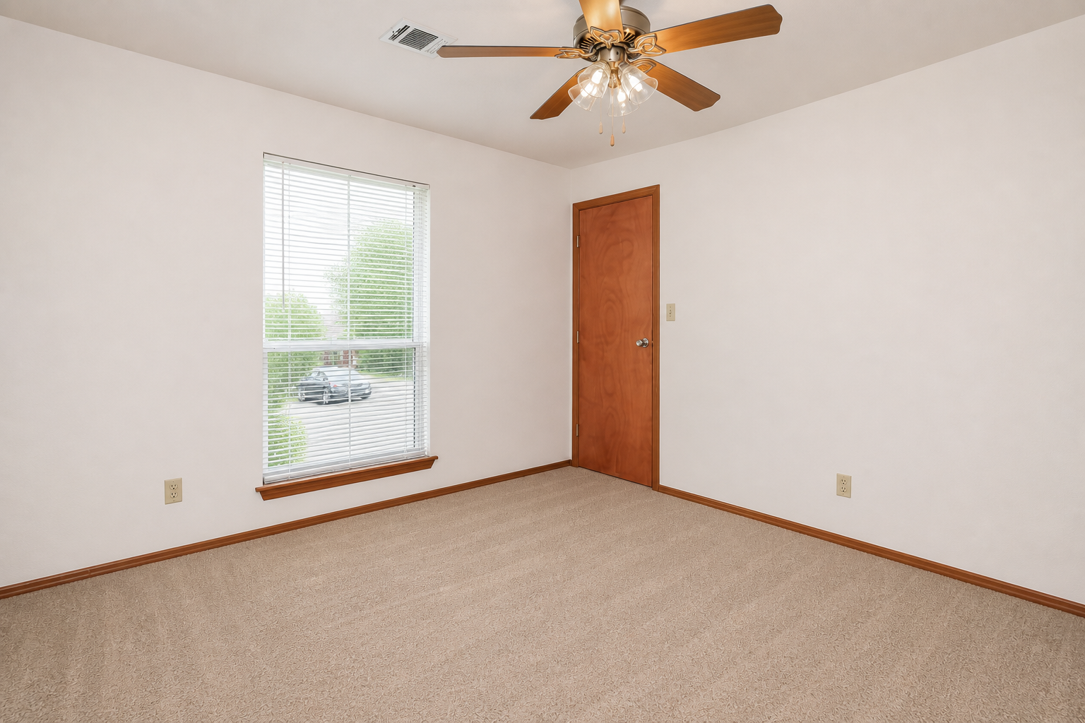 A light-colored room with beige carpet, featuring a window with blinds. A ceiling fan with five blades hangs above, and there's a wooden door on one wall. The room is empty, creating a spacious feel, with a small area visible outside the window. The overall atmosphere is bright and inviting.