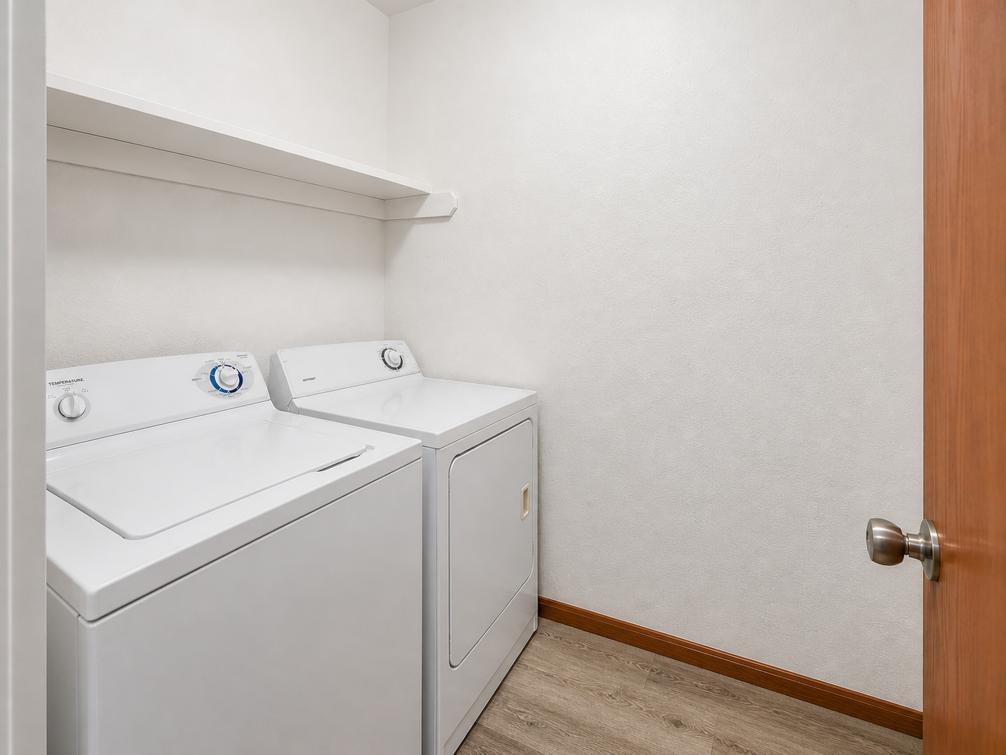 A clean, organized laundry room featuring a white washing machine and dryer side by side, with a shelf above for storage. The walls are light-colored, and there is a wooden door partially open, showcasing a modern and functional space ideal for household chores.