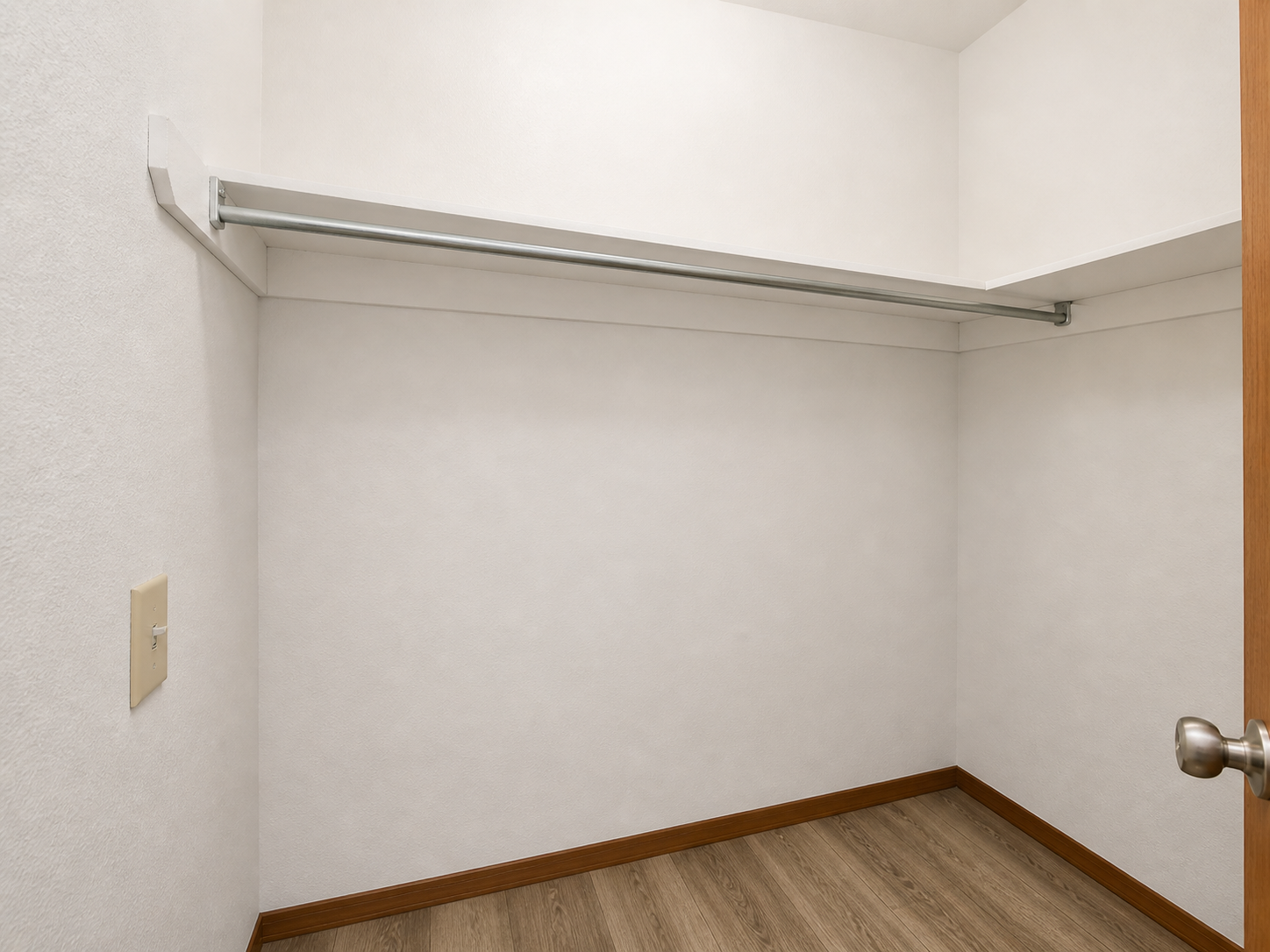 A clean, empty closet with a light-colored wall and wooden floor. It features a metal hanging rod along the top and a door with a brushed metal doorknob. The space is minimalistic and uncluttered, creating a sense of openness.