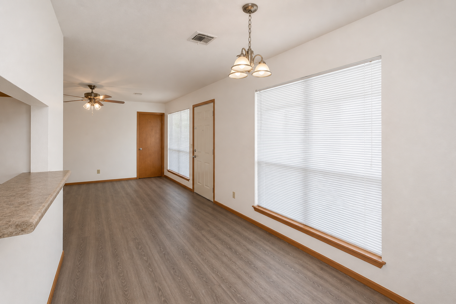 A bright, modern living space featuring light-colored walls and wooden trim. The room has a textured floor, a ceiling fan, and a window with white blinds. A doorway leads to the entrance, and a bar area is visible on the left side. Natural light fills the area, creating an inviting atmosphere.