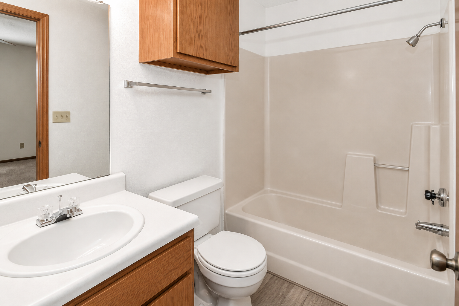 A clean and modern bathroom featuring a white vanity with a sink and mirror, a light-colored bathtub and shower combination, a toilet, and wooden cabinetry. The walls are painted in a neutral tone and the flooring appears to be laminate, contributing to a bright and airy atmosphere.