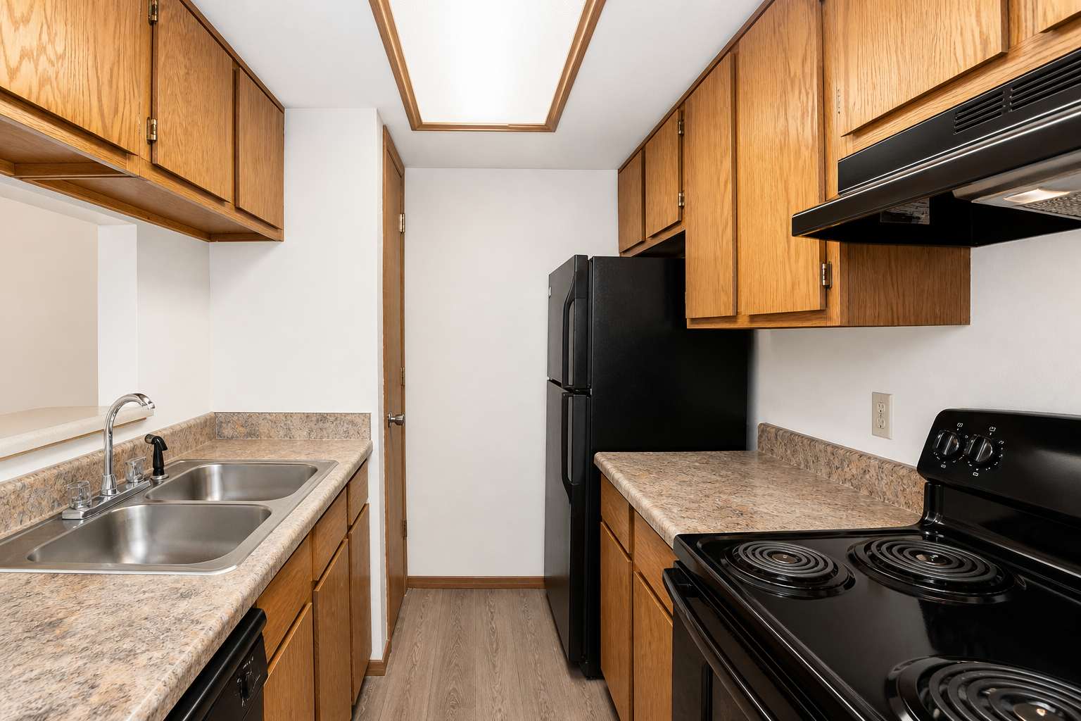 A modern kitchen featuring wooden cabinets, a black refrigerator, and a black stove. The countertop is light-colored with a double sink. The space is well-lit from overhead lighting, and the flooring appears to be a light wood or laminate. The layout is efficient and designed for functionality.