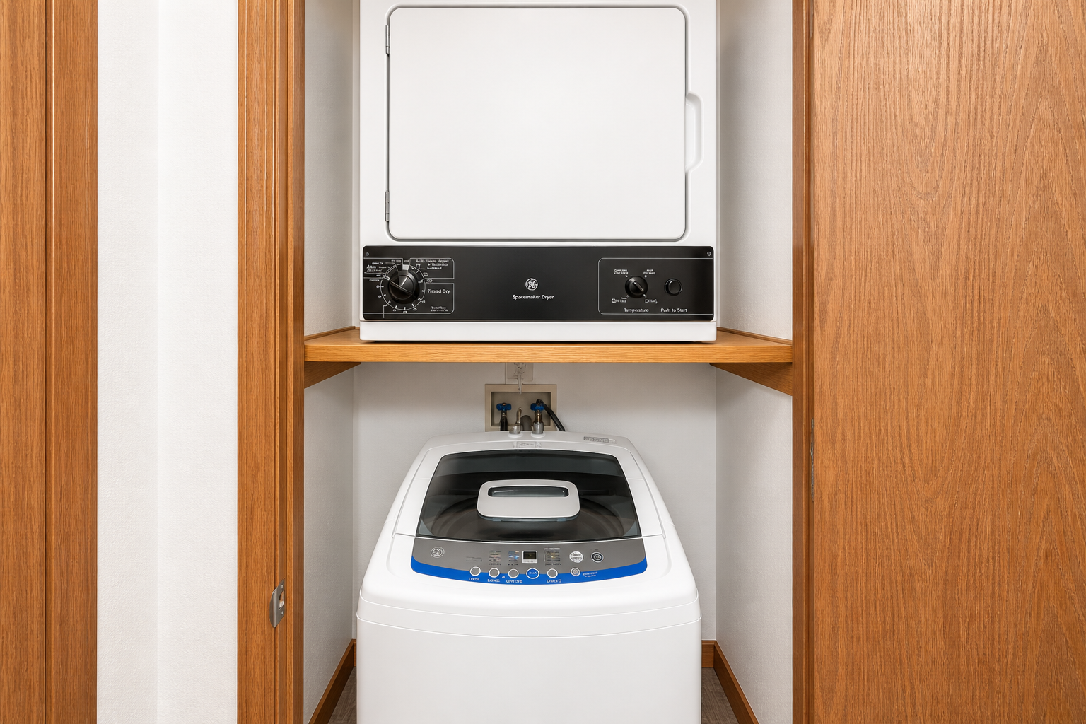 A stacked washer and dryer unit located in a wooden cabinet, with the washer at the bottom featuring control buttons and a blue accent, and the dryer above with a simple control panel. The interior space has light-colored walls and wooden cabinetry, providing a neat and organized laundry area.