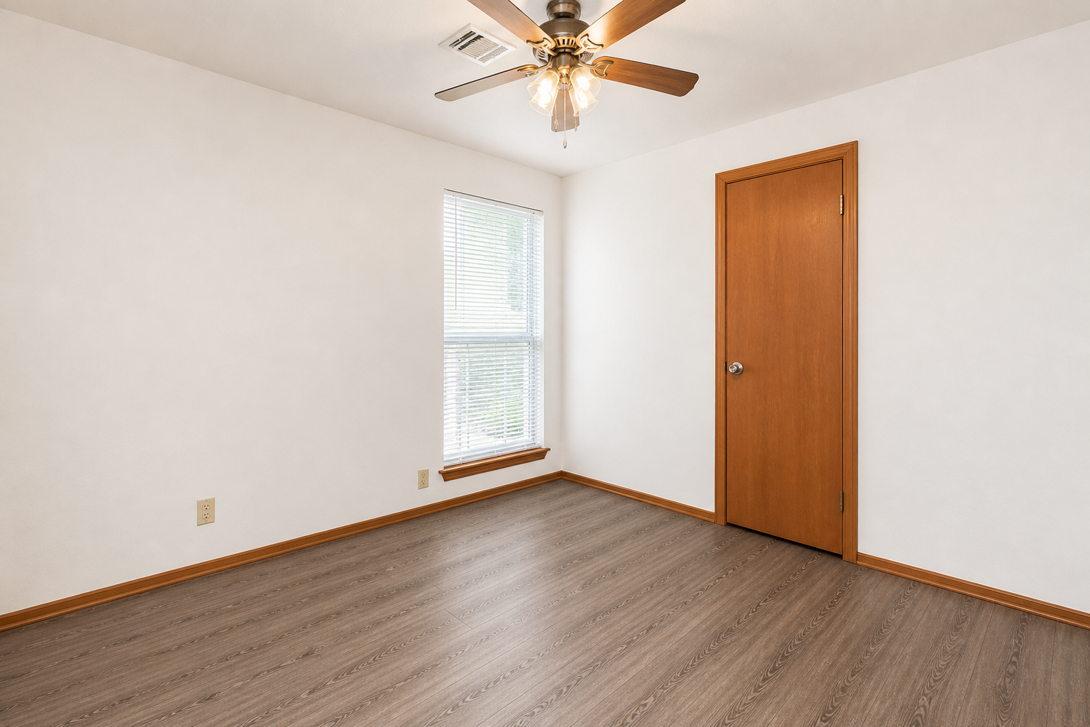 A small, empty room with light-colored walls and a ceiling fan. There is a single window with blinds letting in natural light, and a wooden door on one wall. The flooring is a light wood laminate, and the space has a clean, minimalist aesthetic.
