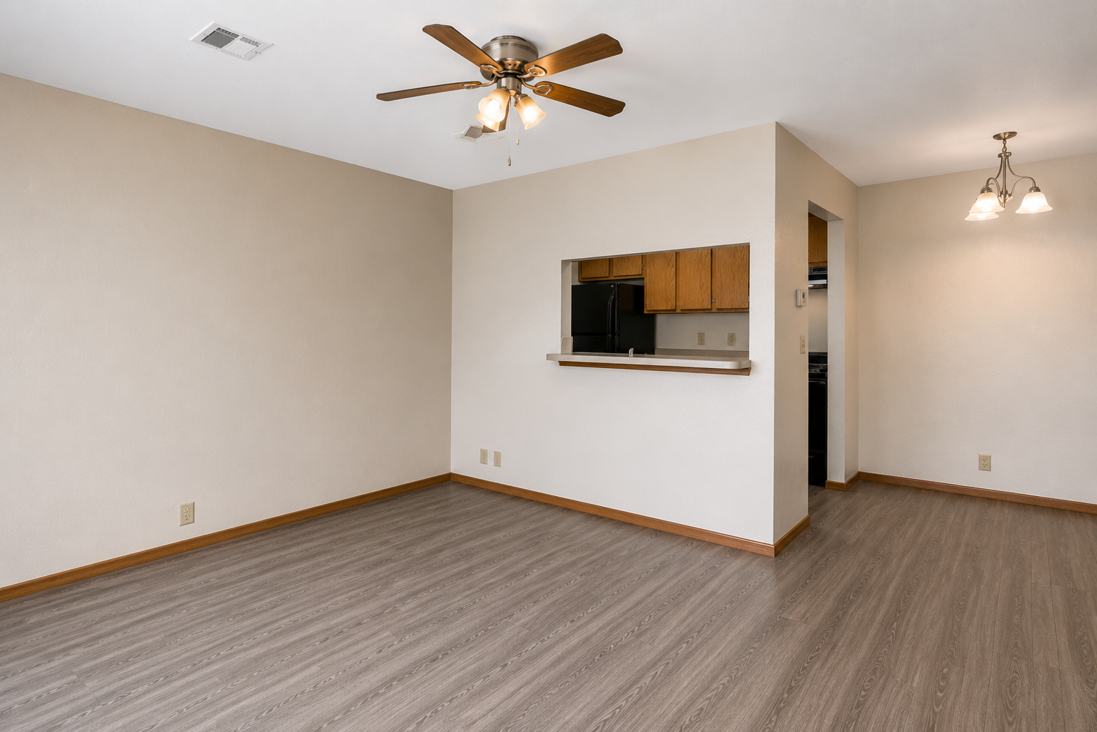 An empty living room with light-colored walls and wood-style flooring. There's a ceiling fan with wooden blades, and an opening to a kitchen area that features wooden cabinetry. Two light fixtures hang above, adding brightness to the space. The area is clean and uncluttered, with no furniture present.