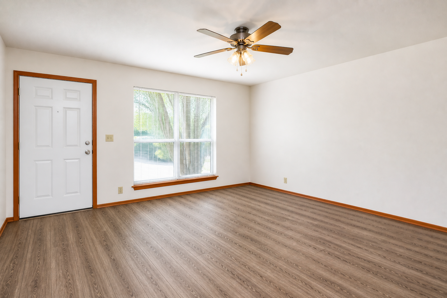 A bright, empty living room featuring a ceiling fan and a large window with vertical blinds. The walls are painted white, and there is light hardwood flooring. A white door is visible on the left side, and the room has a clean, minimalist look, ideal for decorating or furnishing.
