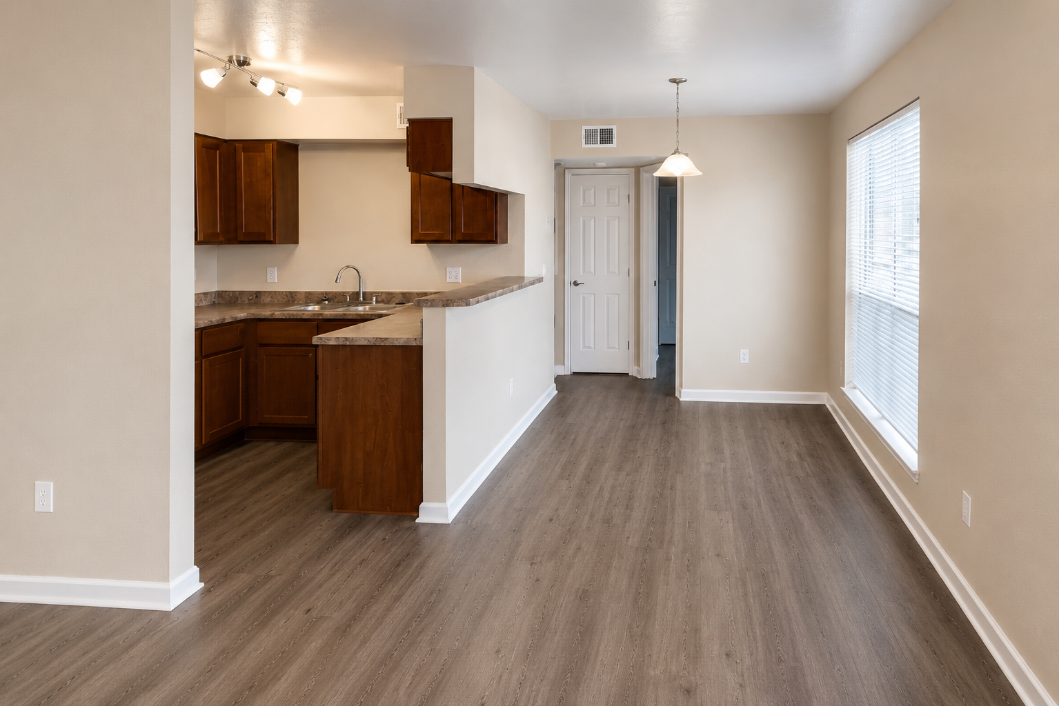 A spacious, modern apartment interior featuring a kitchen with wooden cabinets and a countertop, an open living area with light-colored walls, and a large window allowing natural light. The flooring is a laminate wood design, and there's a door leading to another room.