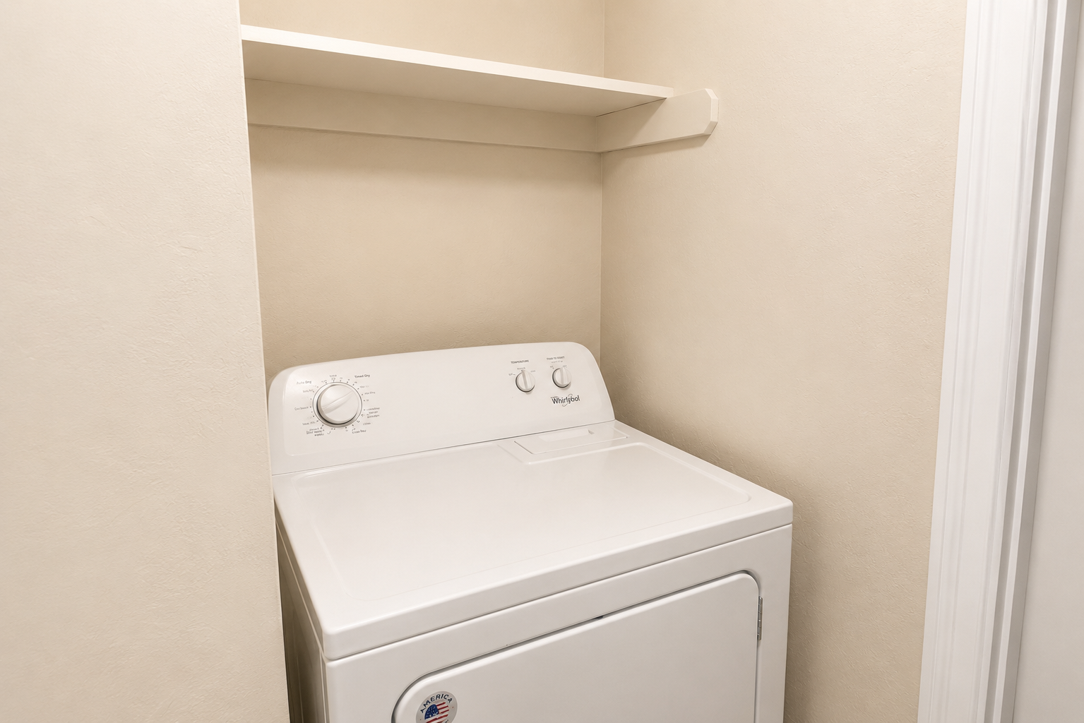 A white top-loading washing machine with a control panel on the front is situated in a small alcove or laundry space. The walls are a light beige color, and there is a simple shelf mounted above the machine, providing storage space.