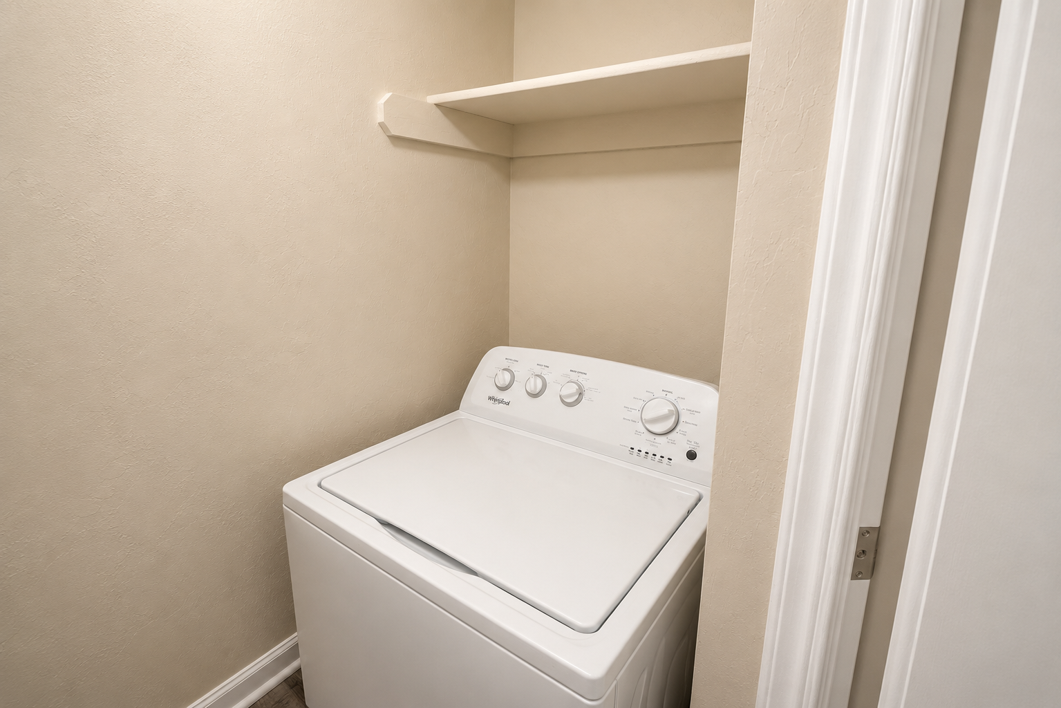 A white washing machine in a small laundry nook with a single shelf above it. The walls are painted in a neutral color, and there is a door frame visible on the right side of the image, suggesting a compact space dedicated for laundry.