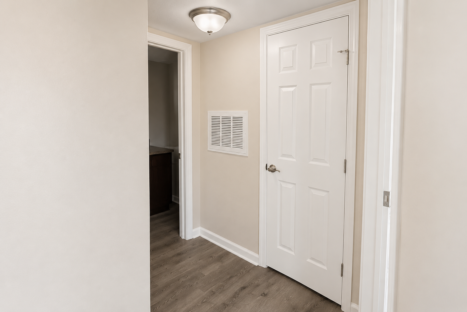 A clean and well-lit hallway featuring light beige walls and a brown wood floor. On the left, there is an open door leading to another room. In the center, a white door with a doorknob and a vent is visible. A ceiling light fixture adds brightness to the space.