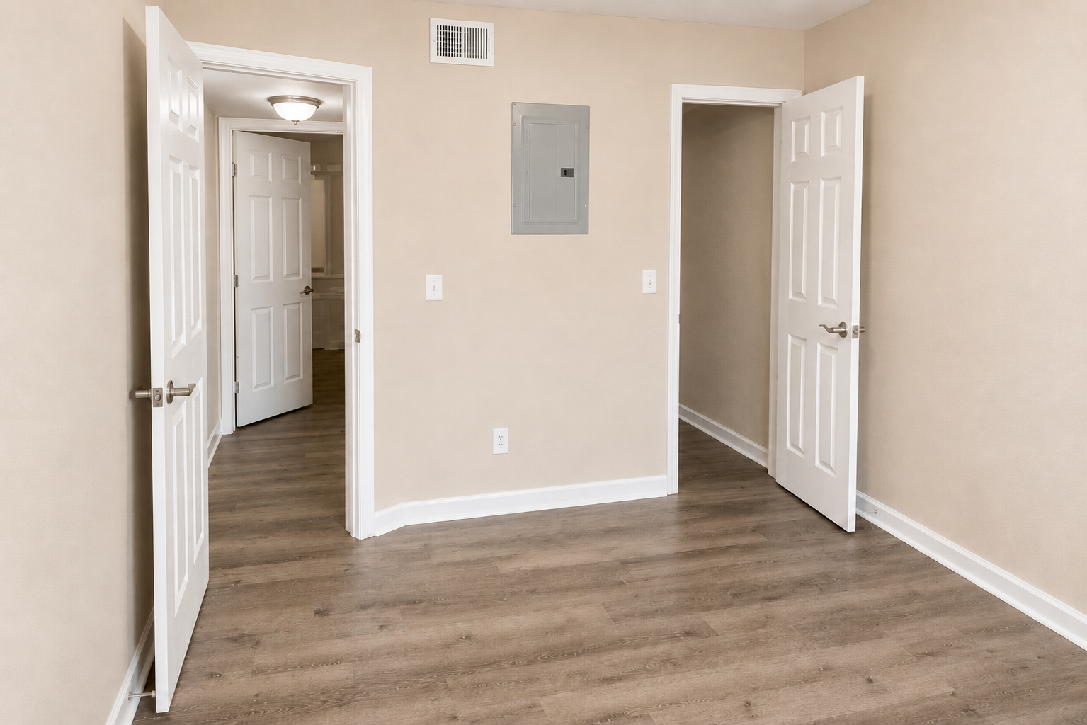 A well-lit, empty room with light beige walls and wooden flooring. There are two open doors on the left and right sides, leading to other rooms. A wall-mounted electrical panel is visible near the center of the room. The space appears clean and ready for furnishing.