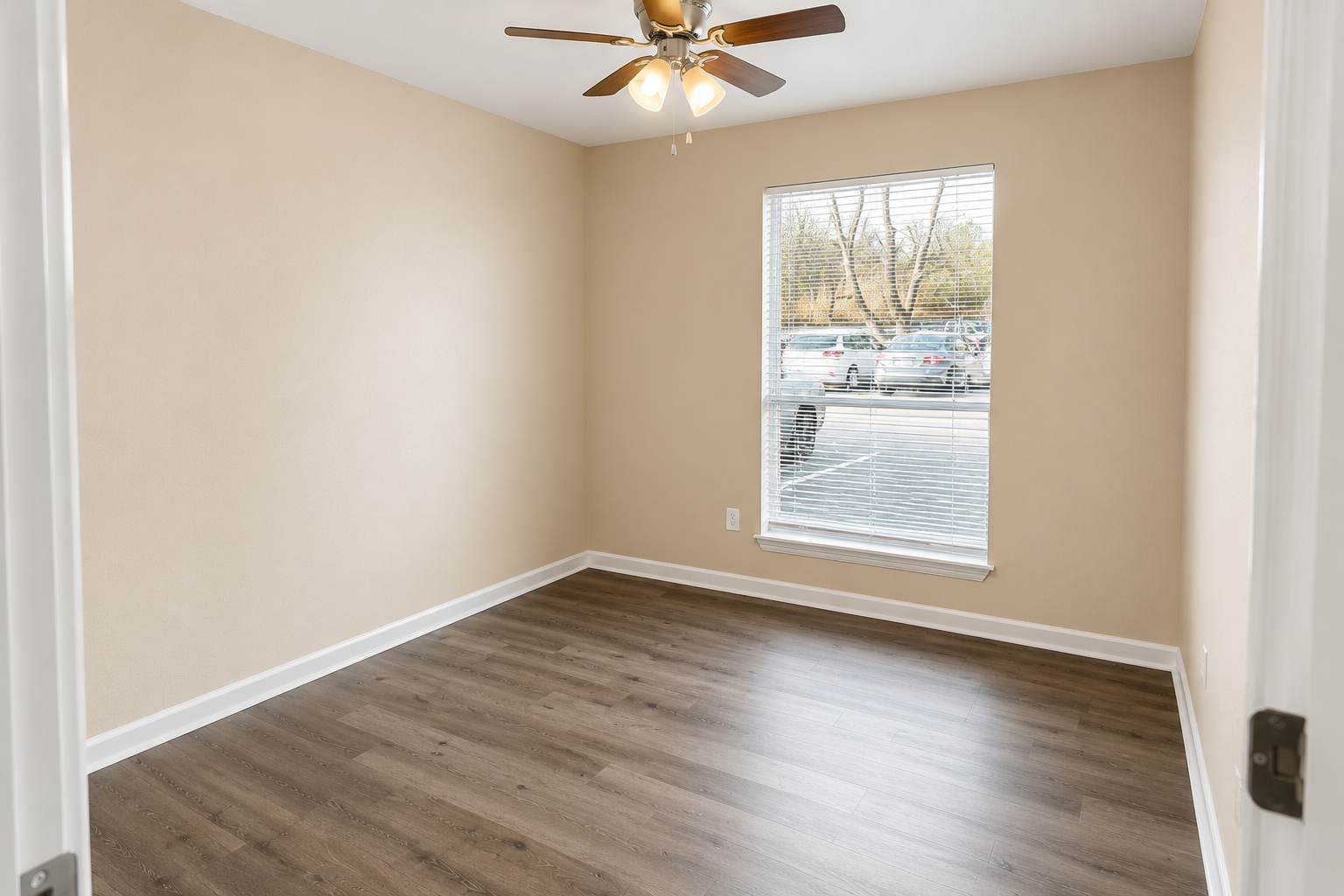A well-lit empty room with light beige walls and a ceiling fan. The room features a window with blinds, offering a view of a parking area outside. The floor has a wood-like finish, and there are no furnishings or decorations, creating a spacious and clean feel.