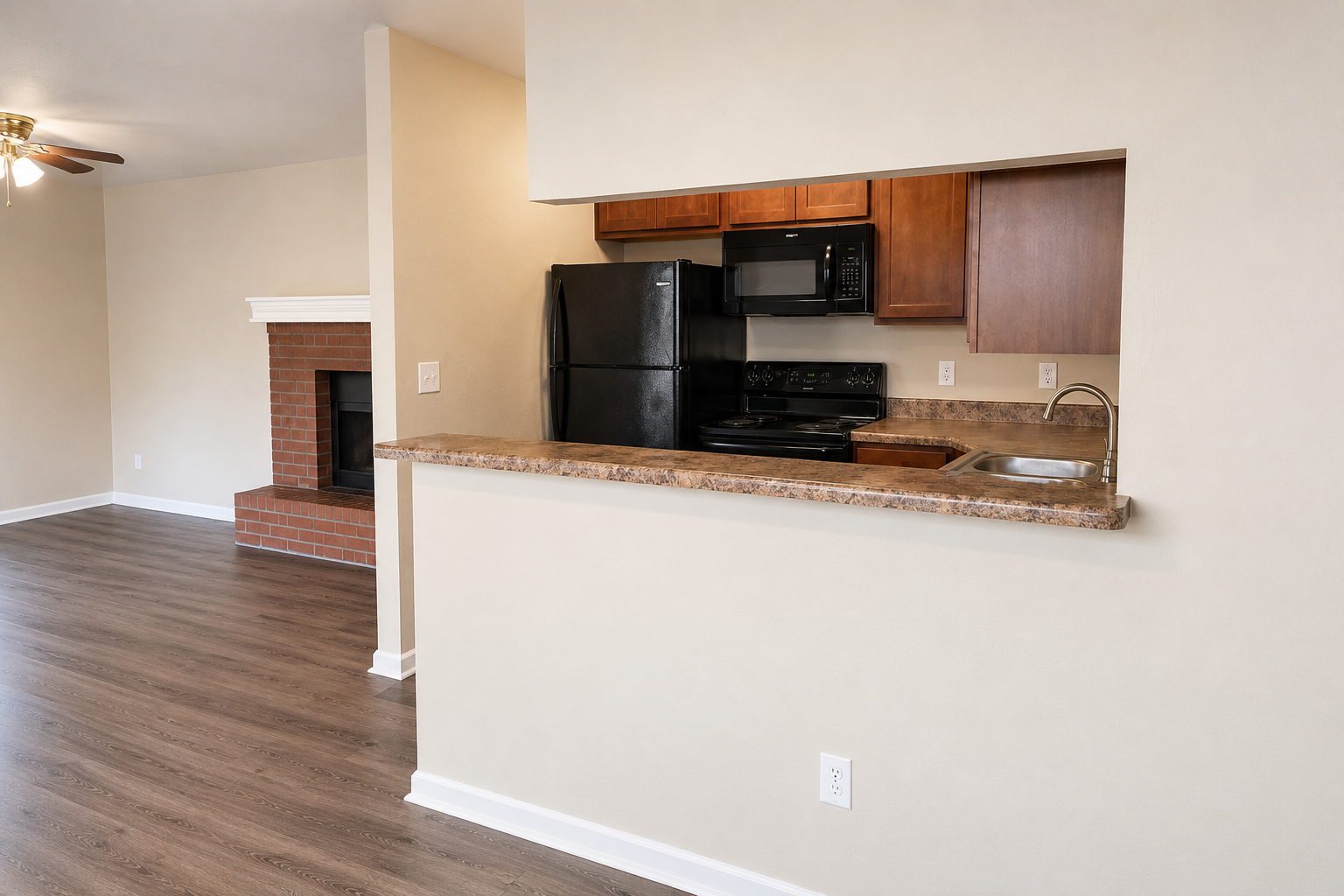 A view of a modern kitchen area with a bar counter opening into a living room. The kitchen features dark wood cabinets, a black refrigerator, and a built-in microwave over the stove. A brick fireplace is visible in the living room, which has hardwood flooring and light-colored walls.