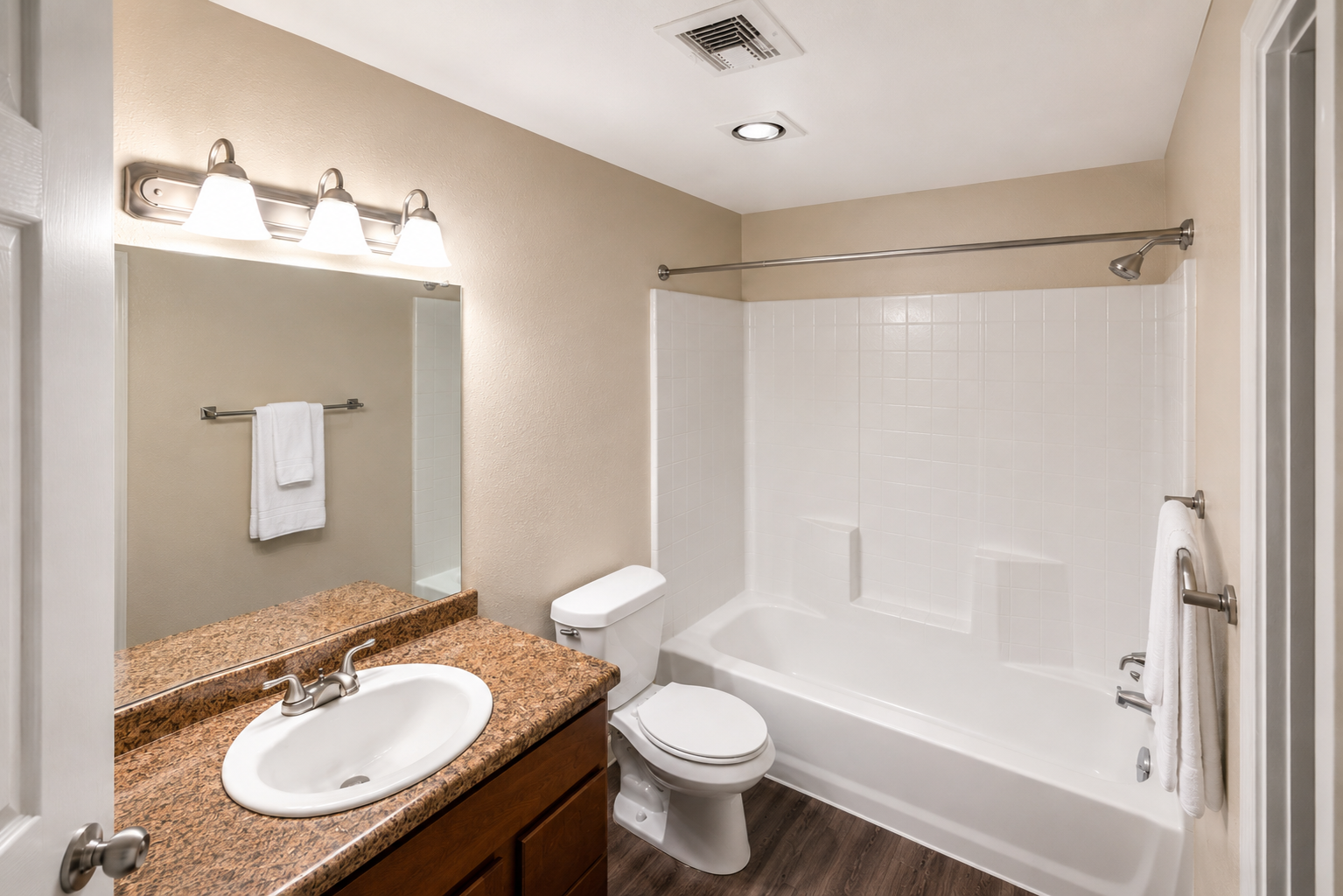 A clean and modern bathroom featuring a white bathtub with a shower, a granite countertop sink with a faucet, a mirror above the sink, and additional towel bars. Soft lighting illuminates the space, which has light-colored walls and a tiled floor.