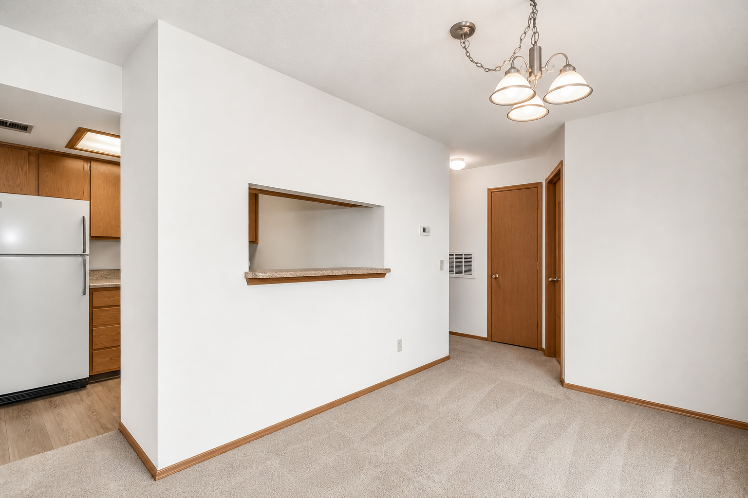 A spacious, well-lit room featuring neutral-colored walls and carpet. There is a small opening, or pass-through, to a kitchen on the left with wooden cabinets and a white refrigerator. The ceiling displays a modern light fixture with three bulbs, and there is a door on the right leading to another room.