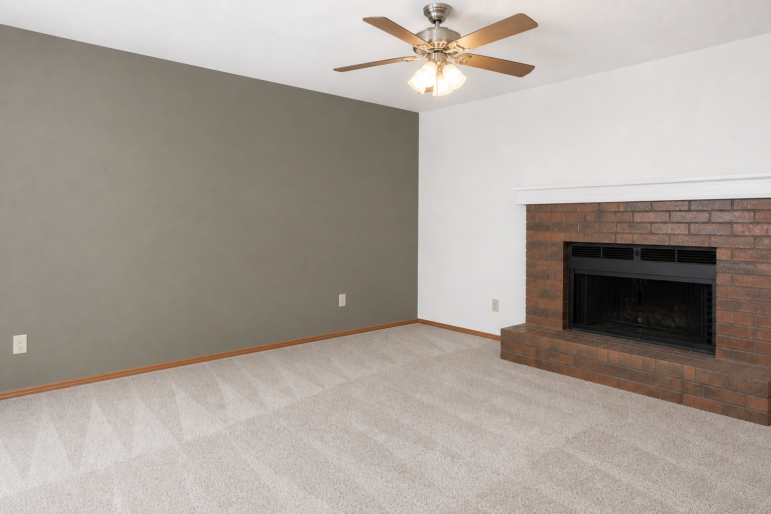 A spacious living room featuring a brown brick fireplace, a ceiling fan with four lights, and light beige carpet. One wall is painted a muted green, while the opposite wall is white. The room is empty, providing a clean and inviting atmosphere suitable for various furnishings and decorations.