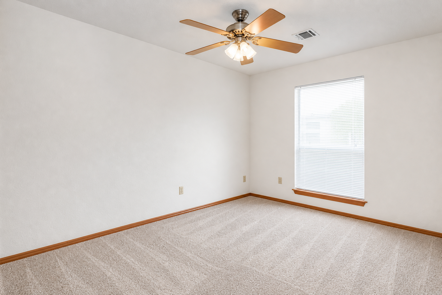 A bright, empty room featuring light beige walls and a light brown carpet. The room is illuminated by a ceiling fan with a light fixture. There is one window with blinds, allowing natural light to enter. The space is uncluttered and ready for furniture placement.