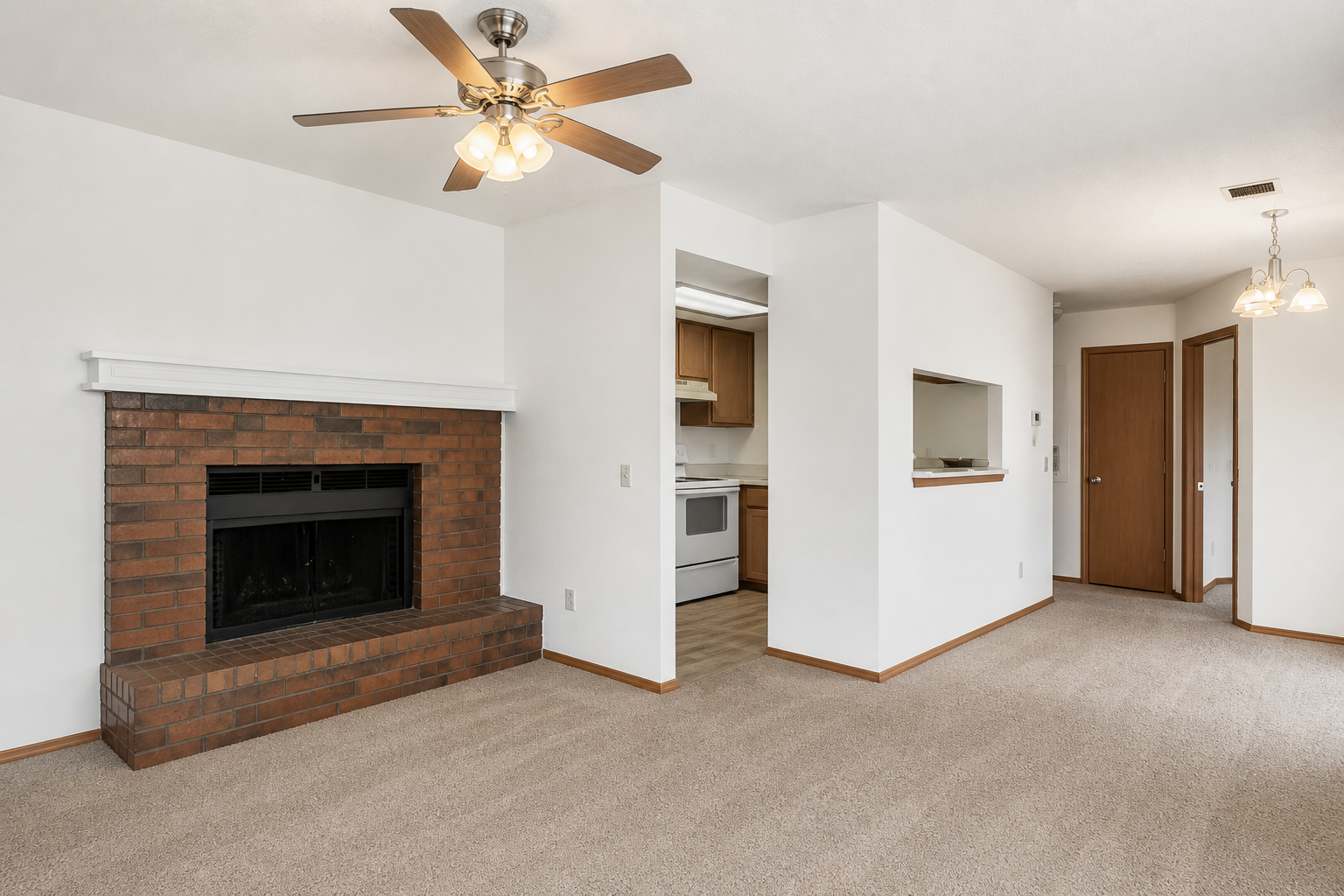 Living room with beige carpet and white walls. A brick fireplace is on the left with a ceiling fan above. To the right, there is an open kitchen area with wooden cabinets and a doorway leading to another room. Soft lighting fixtures hang from the ceiling, contributing to a cozy atmosphere.