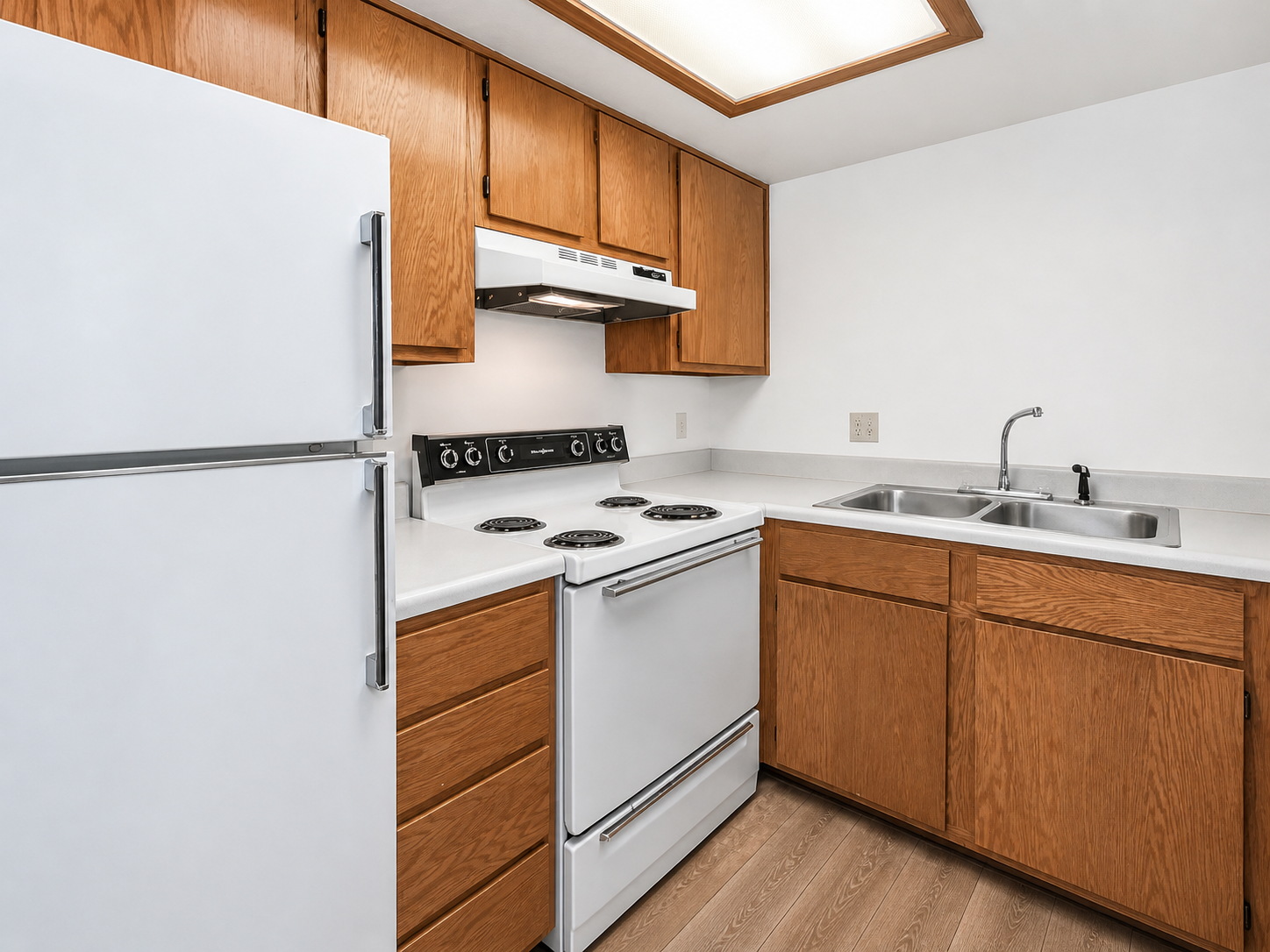 A modern kitchen featuring light wood cabinets, a white refrigerator, an electric stove with an overhead microwave, and a stainless steel sink. The countertops are light-colored, and the flooring appears to be laminate with a light wood finish. The space is bright and tidy, with a simple, functional layout.