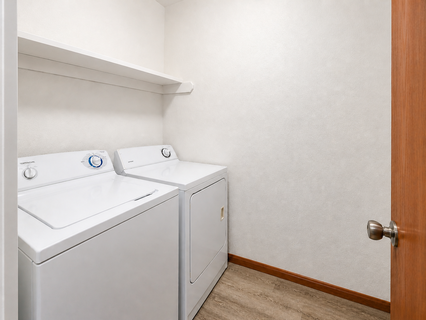 A laundry room featuring two white washing machines side by side against a light-colored wall. There is a shelf above the machines, and a wooden door partially visible on the right. The floor is a light, wood-like material.