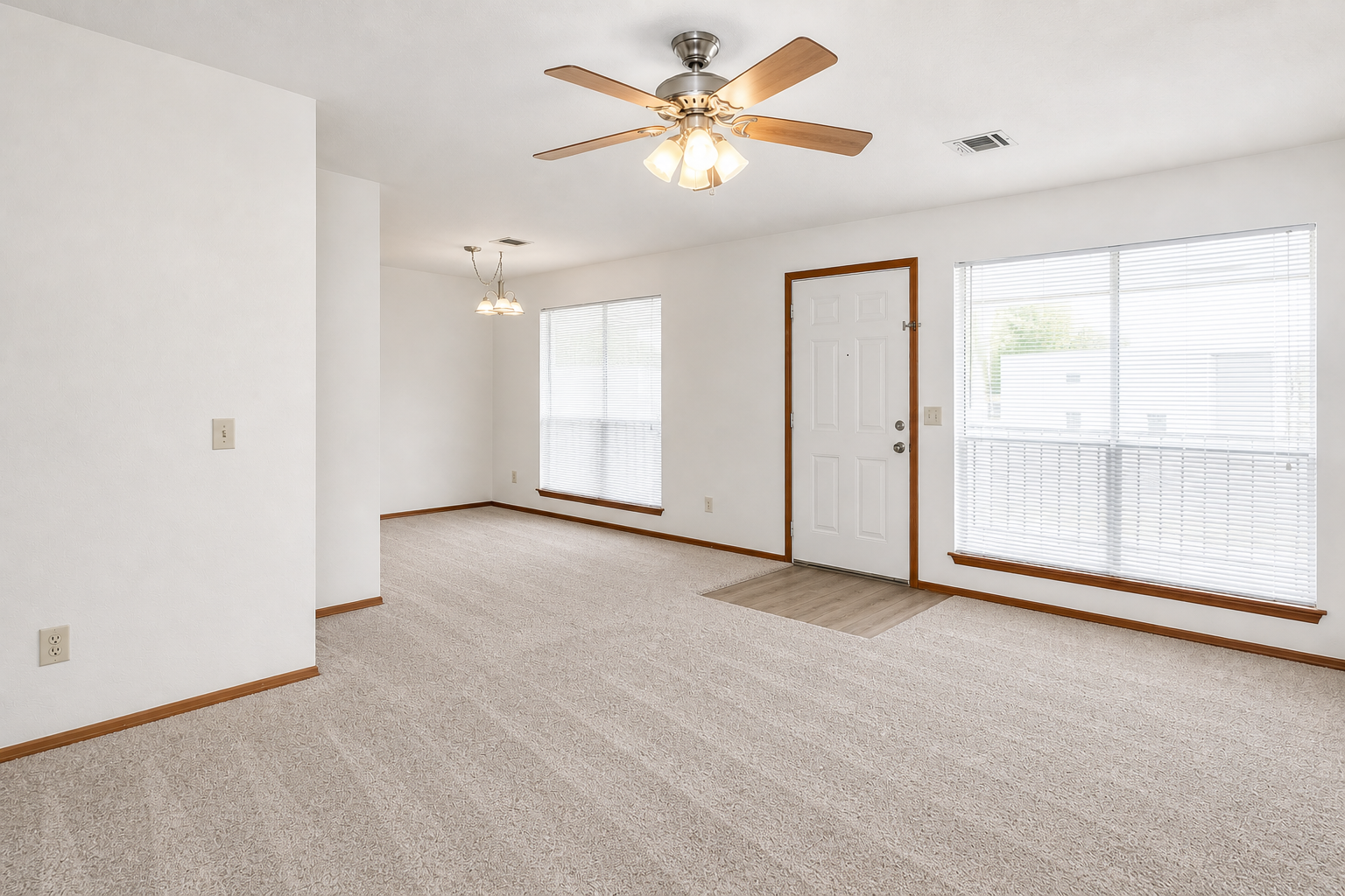 Spacious, light-filled living area featuring neutral-colored walls and plush carpet. Two large windows allow natural light to flood the room, complemented by a ceiling fan. A front door with a small entryway mat leads outside, while a dining area with a hanging light fixture is visible in the background.