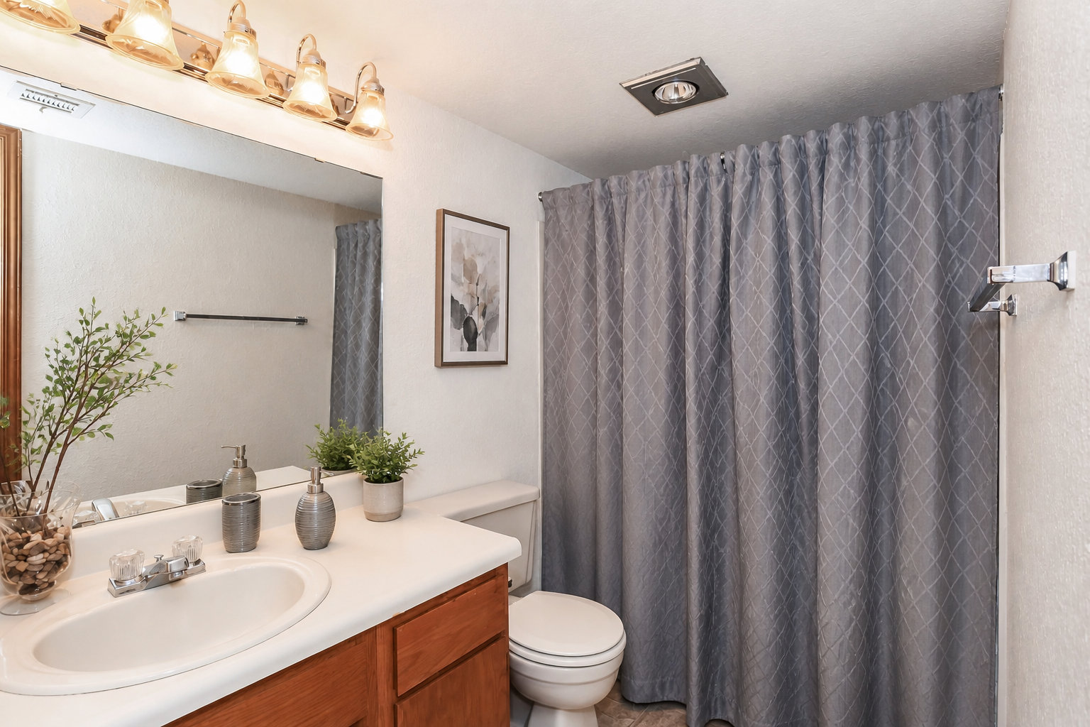 A clean and modern bathroom featuring a light-colored countertop, wooden cabinetry, and a decorative plant. The space includes a white toilet, a shower with a grey curtain, and a framed artwork on the wall. Soft lighting is provided by four overhead fixtures.