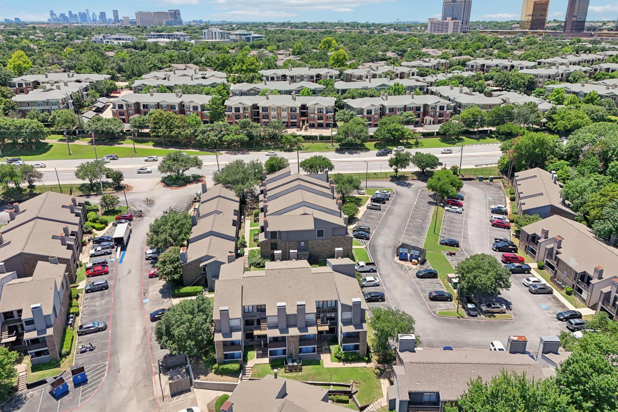 Aerial view of a suburban area featuring residential buildings, parking lots, and green trees. In the background, a city skyline is visible, indicating proximity to urban development. The scene captures a mix of apartments and townhouses, with streets and vehicles throughout the neighborhood.