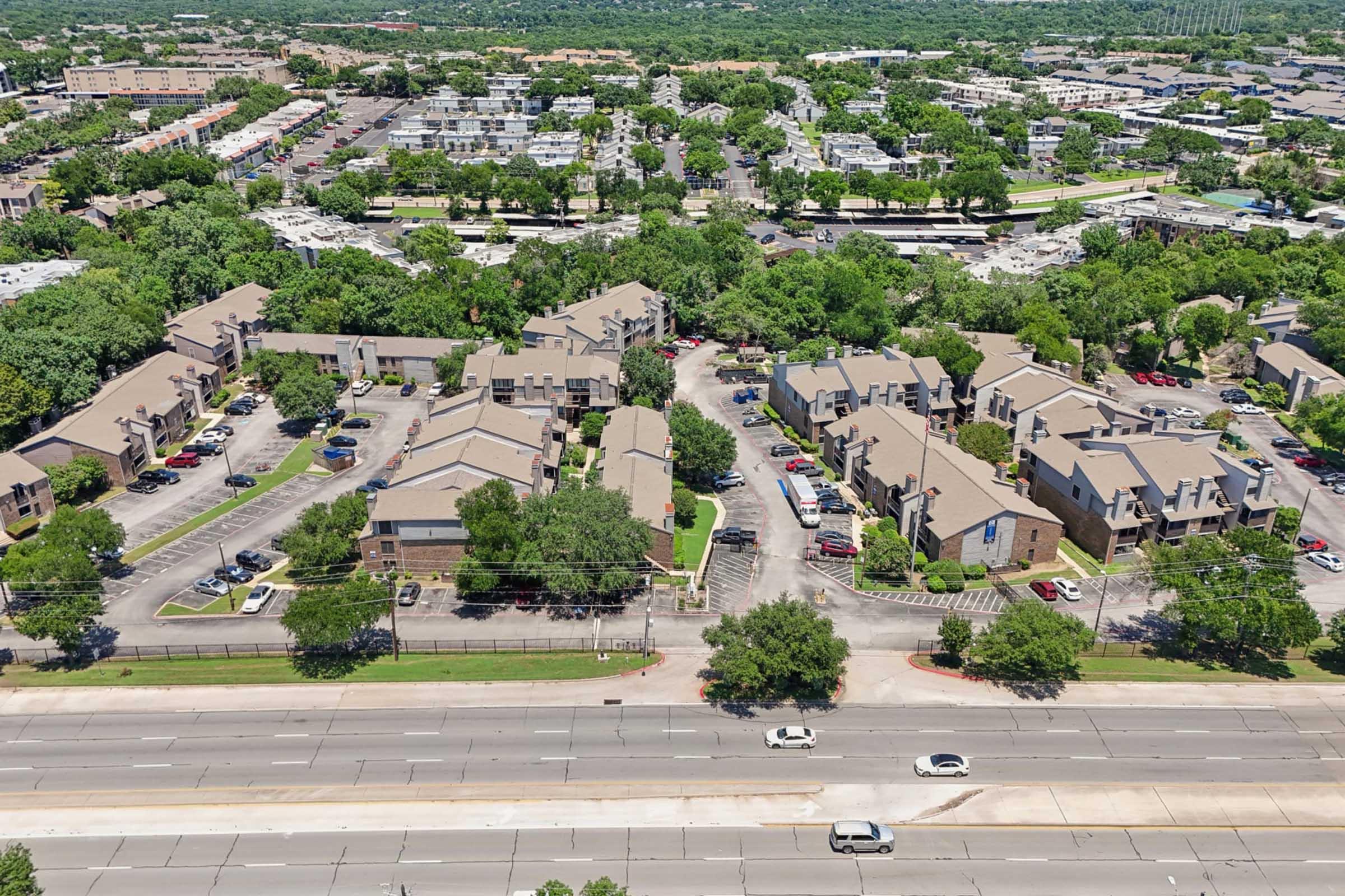 Aerial view of a residential area featuring multiple apartment buildings surrounded by trees and greenery. Roads and parking lots are visible, with several cars parked. In the background, more buildings and urban development can be seen, indicating a suburban environment near a highway.