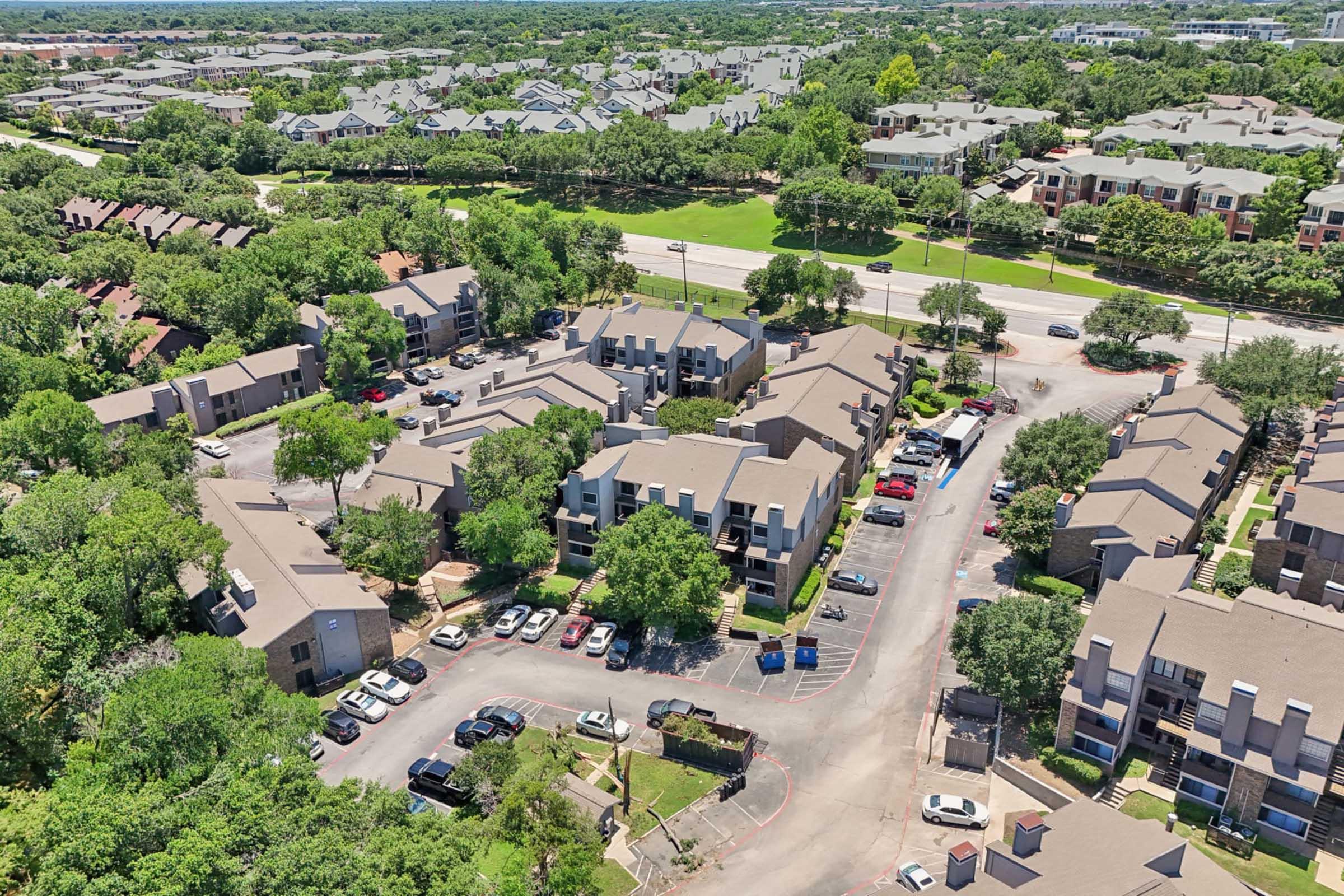Aerial view of a residential complex featuring multiple apartment buildings surrounded by trees and parked cars. A road runs along the side of the community, with additional housing visible in the background. The area appears green and well-maintained, suggesting a suburban environment.