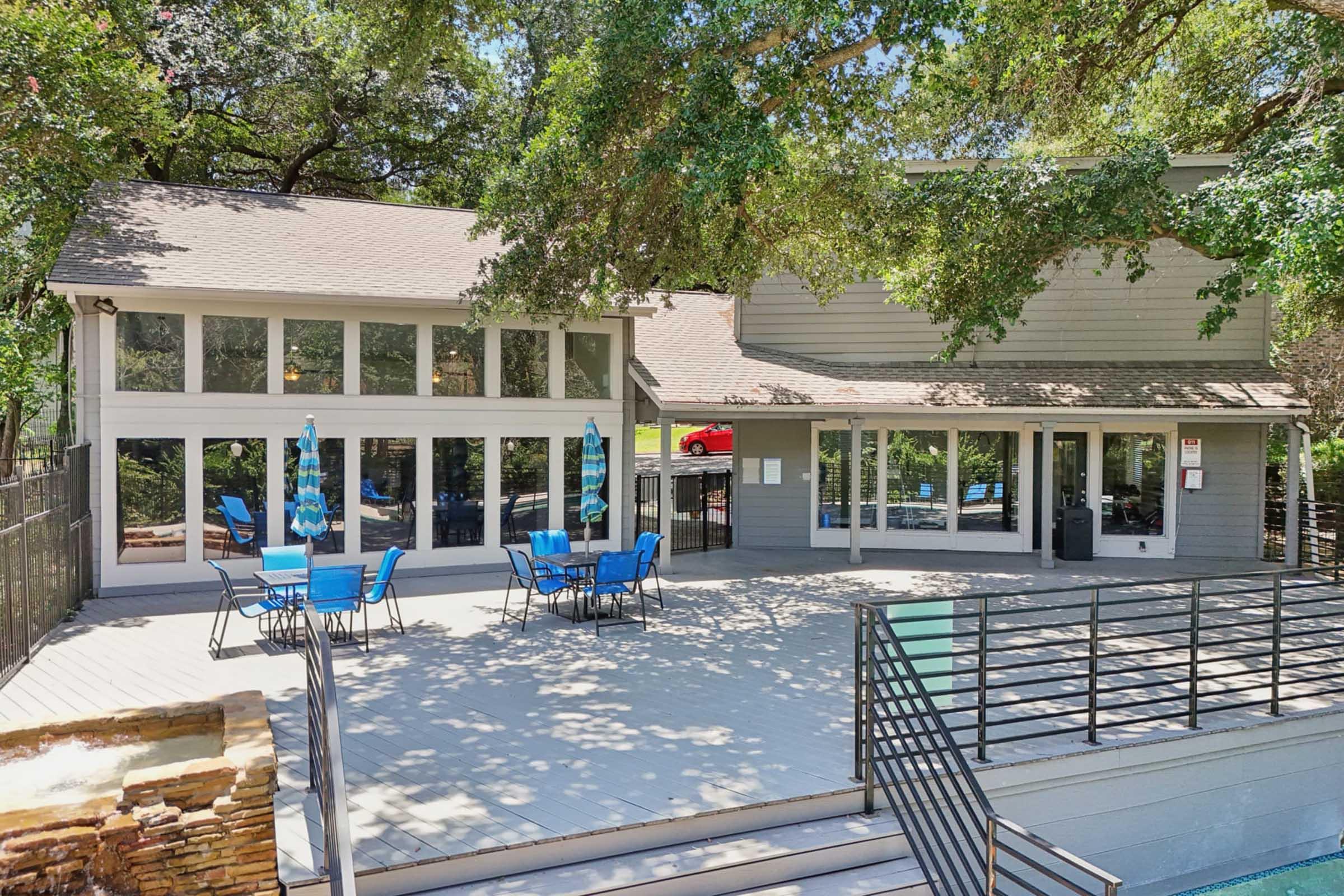 A modern building with a spacious outdoor deck featuring blue chairs and tables. Surrounded by trees, the structure has large windows allowing natural light. A pool area is visible in the foreground, creating a relaxing environment ideal for gatherings or leisure activities.