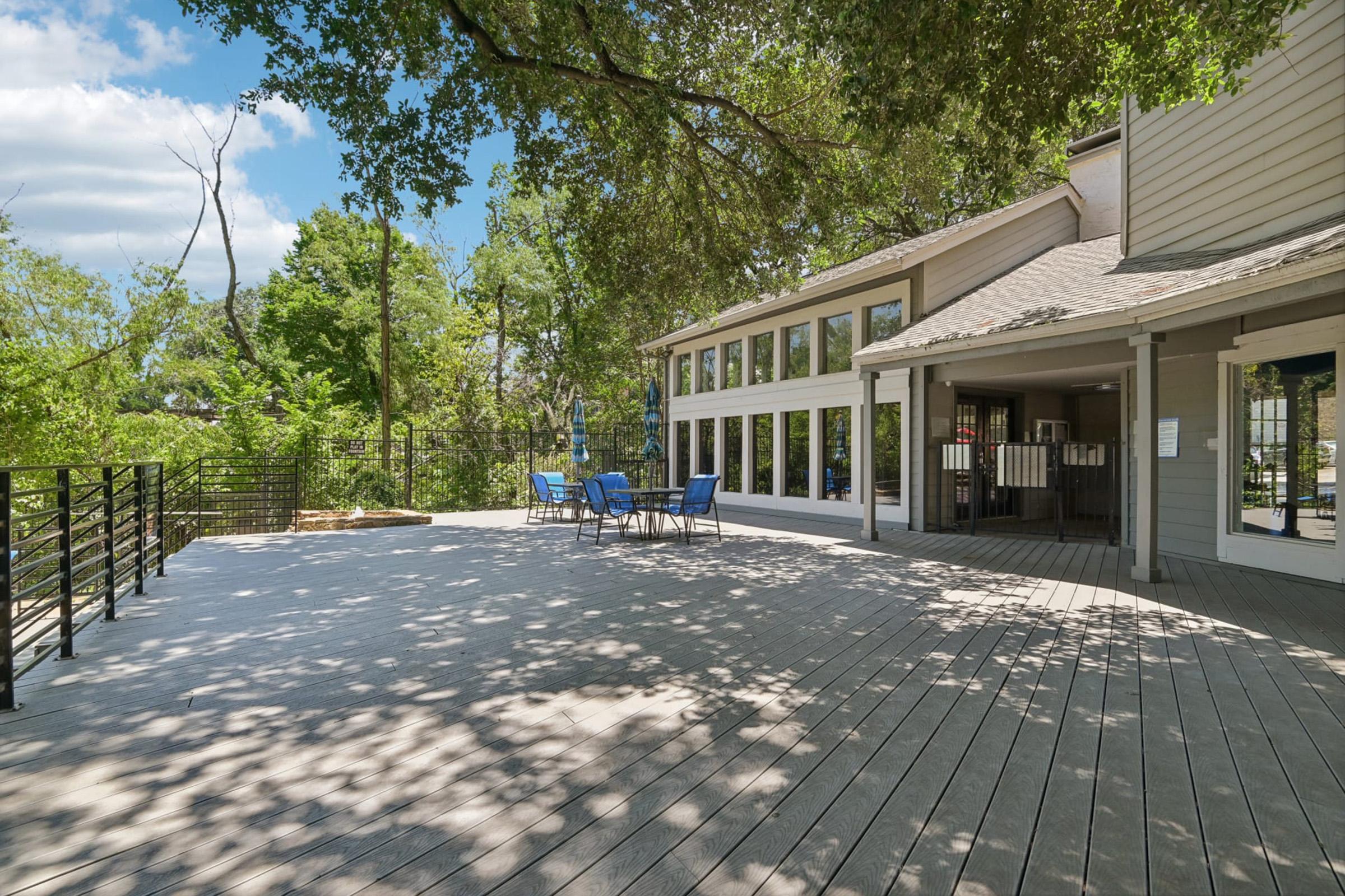 A spacious outdoor deck with blue chairs, surrounded by lush greenery and trees. The deck is attached to a modern building featuring large windows. Soft sunlight filters through the leaves, creating a peaceful and inviting atmosphere, ideal for relaxation or gatherings.
