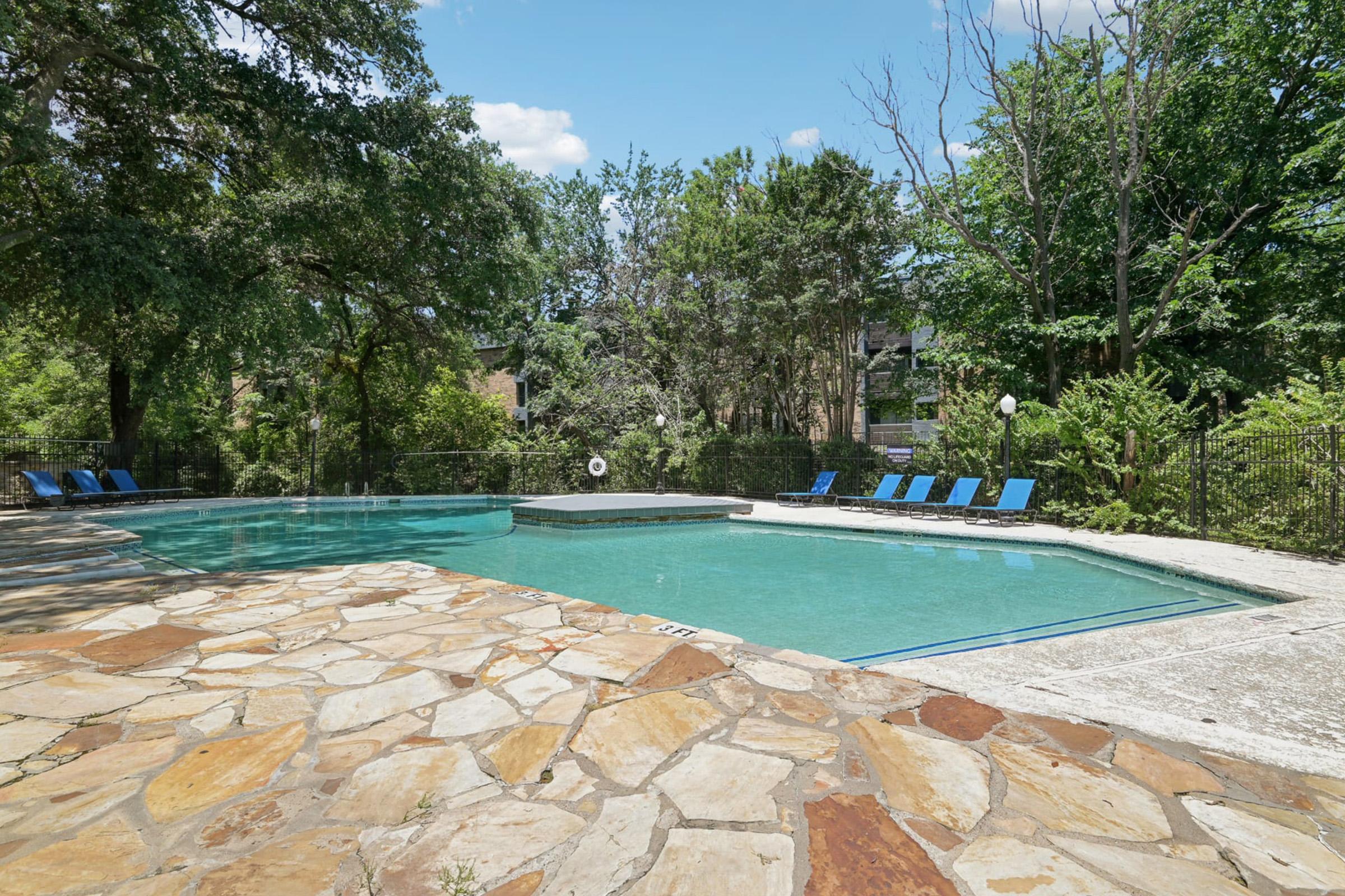 A serene outdoor swimming pool surrounded by lush greenery, featuring blue lounge chairs along the edges. The pool area is framed by natural stone and shaded by trees, creating a relaxing atmosphere on a sunny day.