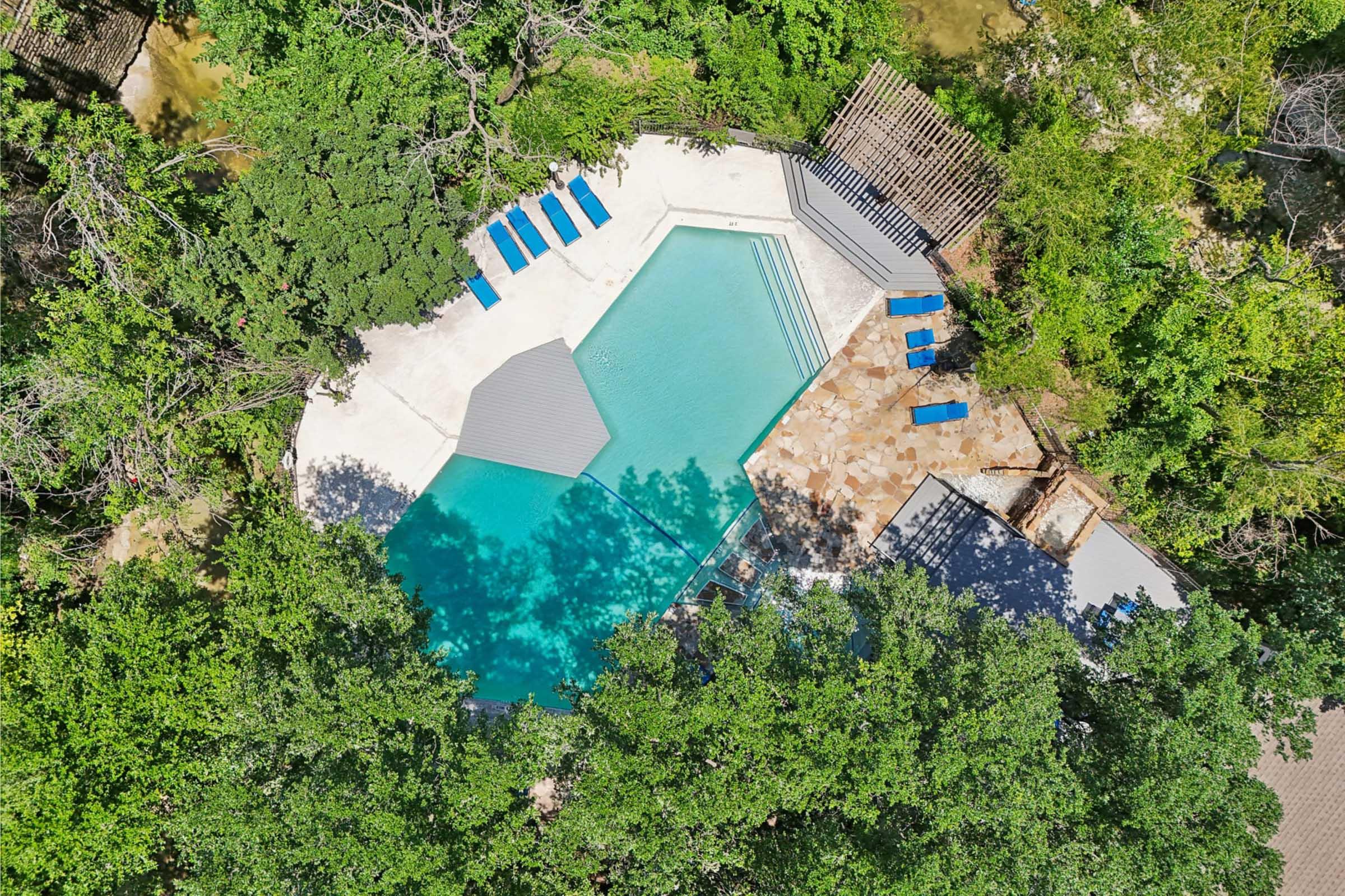 Aerial view of a modern swimming pool surrounded by lush greenery. The pool features an interesting geometric shape, with a deck area and several blue lounge chairs arranged nearby. Natural stone and wooden elements create a serene outdoor environment.