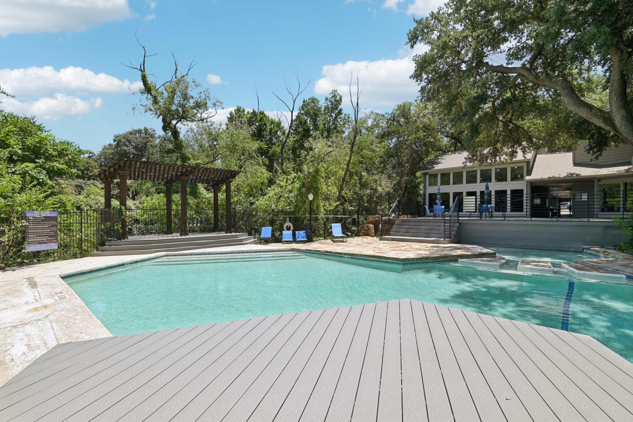 A clear swimming pool surrounded by a wooden deck, with lounge chairs nearby. Lush greenery and trees are visible in the background, along with a house featuring large windows. A pergola stands to the left of the pool, providing a shaded area. The sky is partly cloudy, giving a serene atmosphere.
