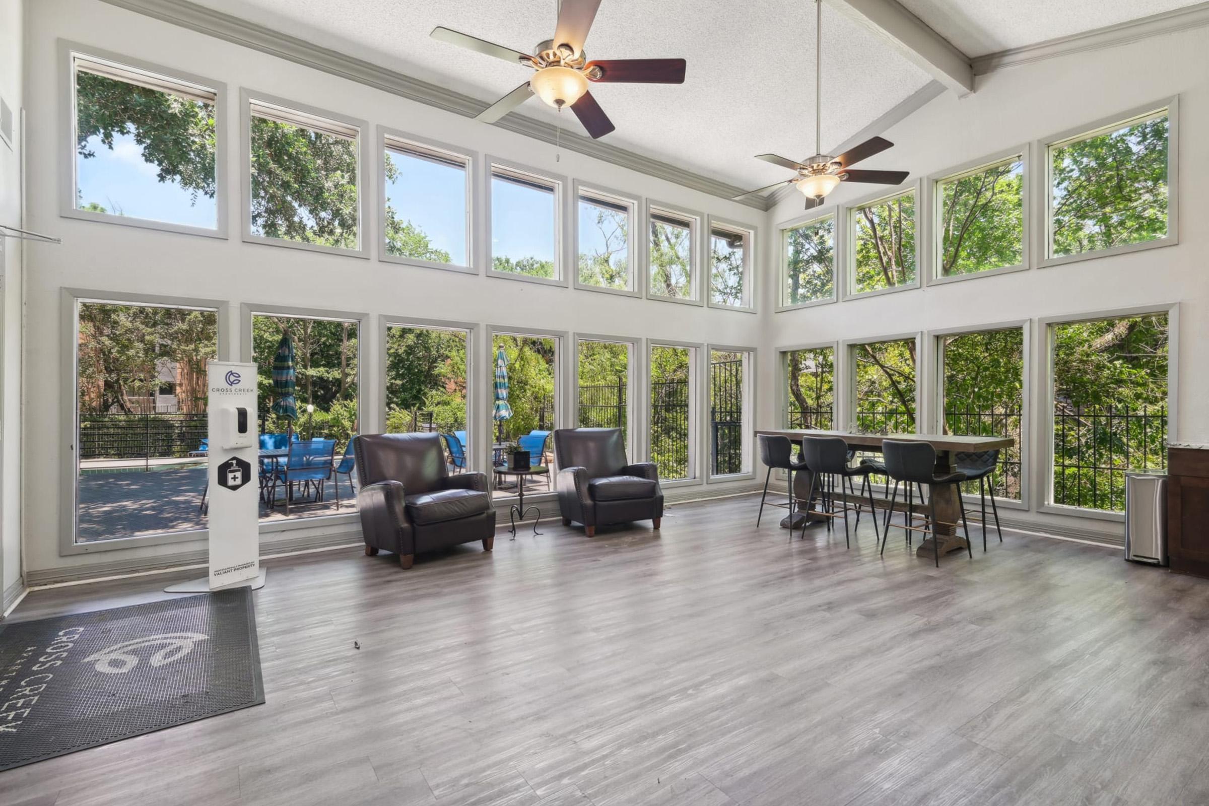 Bright and airy common area featuring large windows that let in natural light, two comfortable chairs, a modern table for seating, and a sleek charging station. The view outside includes greenery and outdoor seating options. The space has a contemporary design with high ceilings and a welcoming atmosphere.