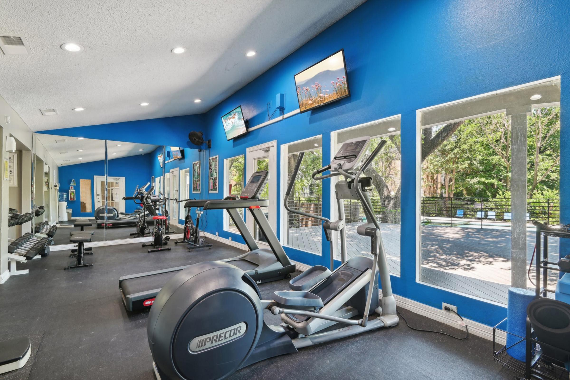 A modern gym interior featuring exercise equipment like treadmills and weights, with large windows allowing natural light. The walls are painted blue, and there are televisions mounted on the walls, creating a vibrant and inviting workout space.