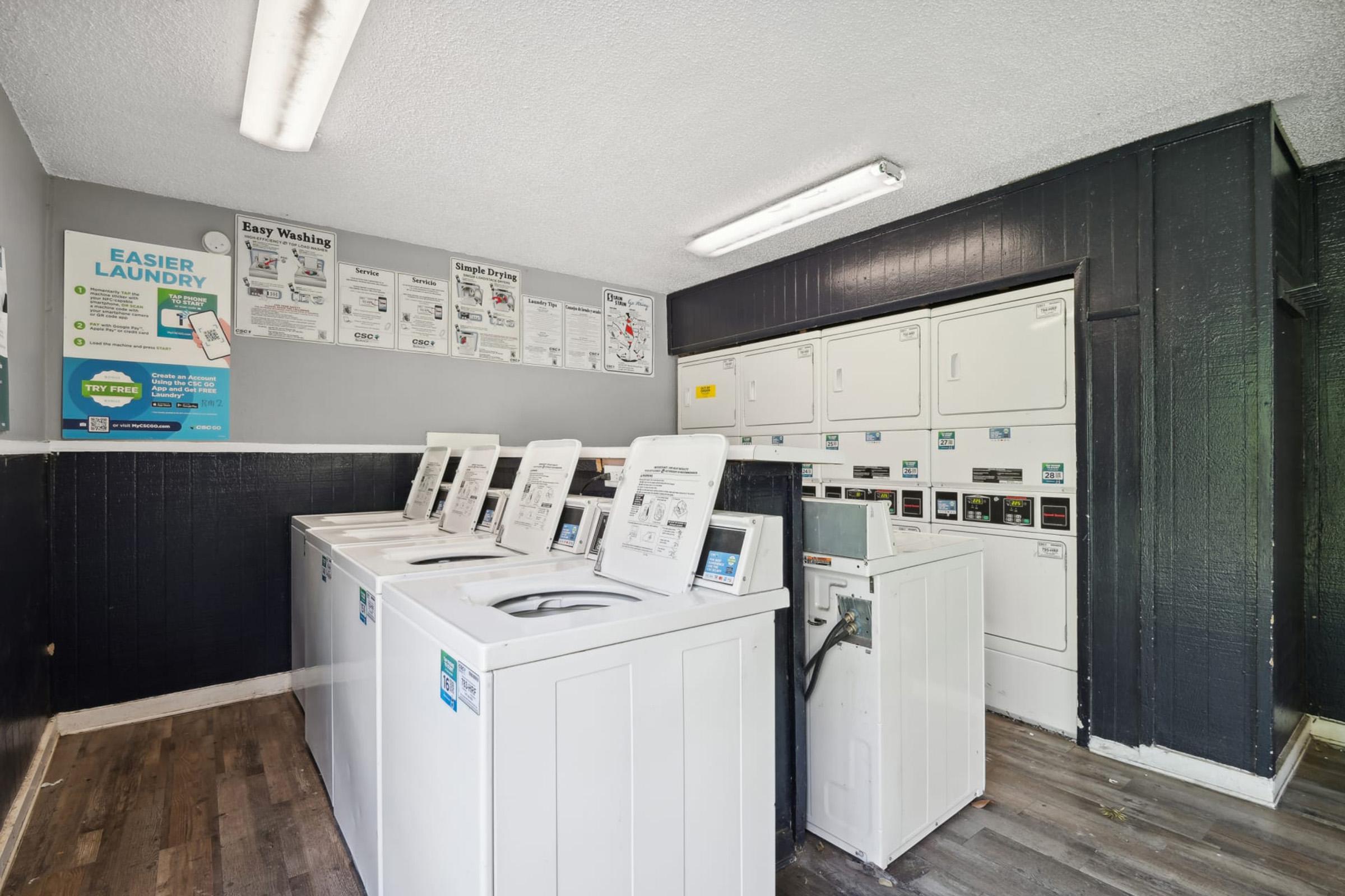 A clean and well-lit laundry room featuring multiple white washing machines and dryers. The walls are adorned with informational posters about laundry services, while the flooring is a wooden laminate. The space is organized, providing a practical environment for laundry tasks.