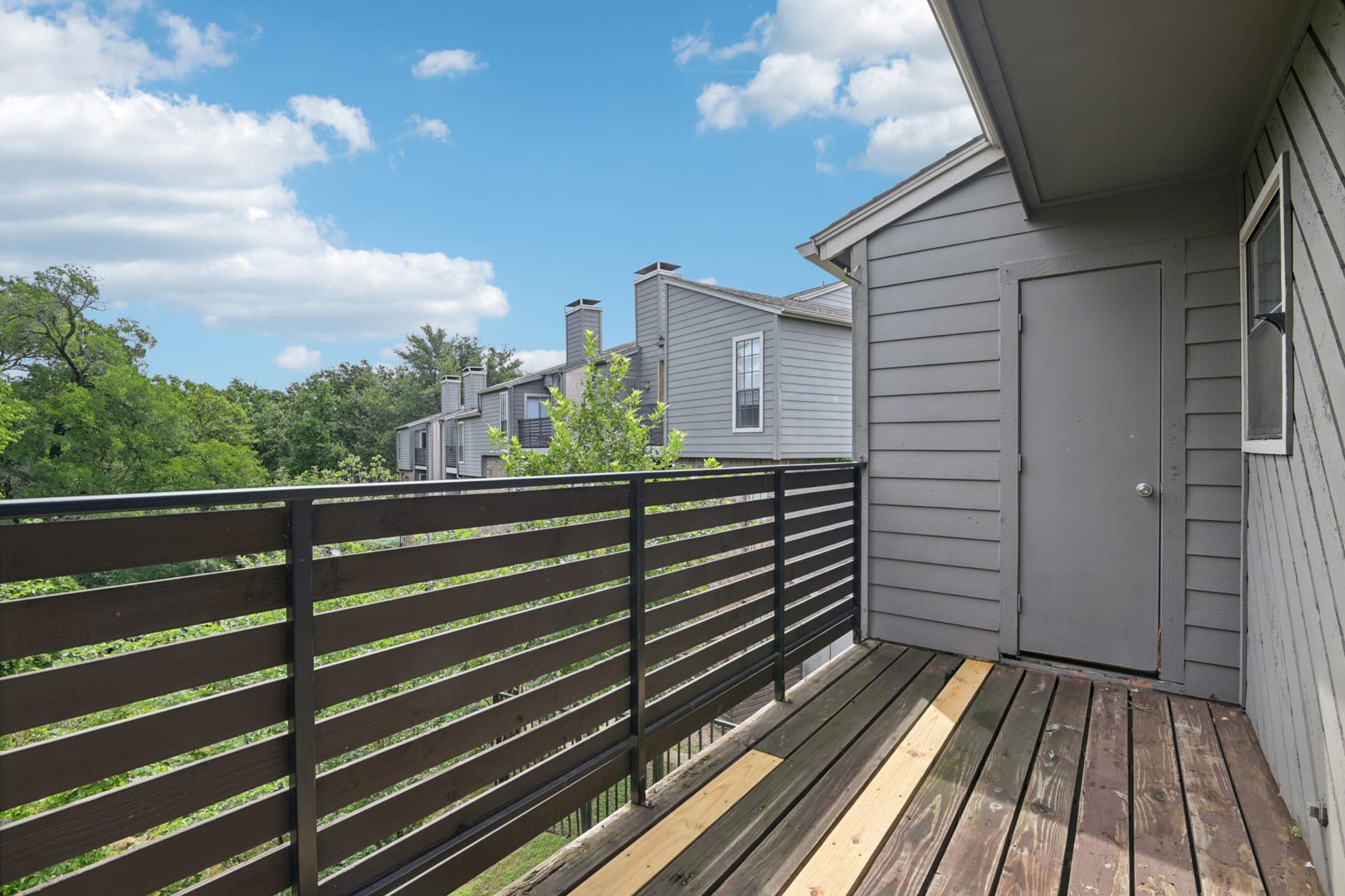 A balcony view featuring a wooden deck with black railing, overlooking green trees and nearby buildings. The sky is partly cloudy, adding a bright, open feel to the scene.