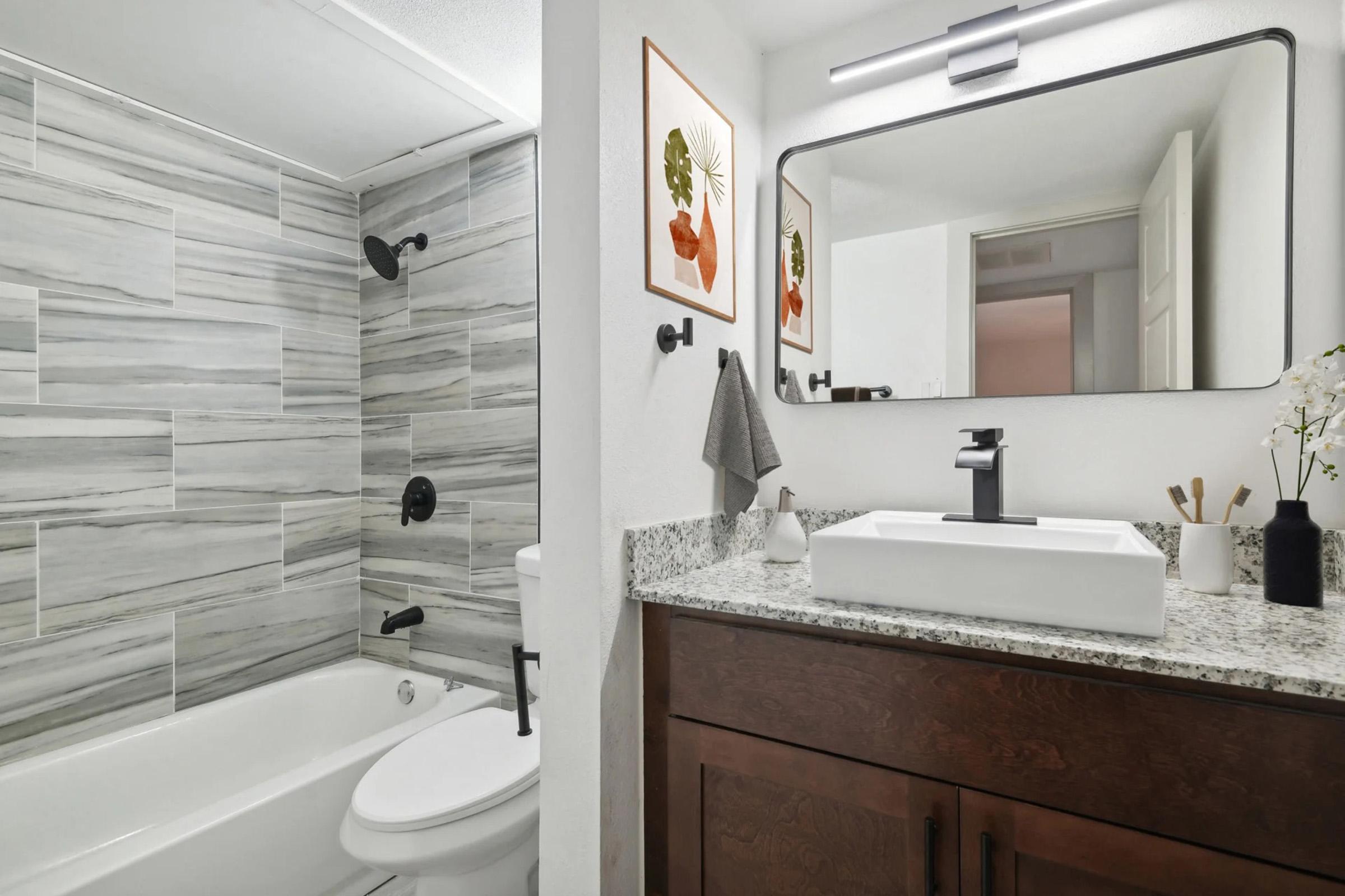 Modern bathroom featuring a white bathtub, sleek gray tiled walls, black fixtures, and a dark wood vanity with a granite countertop. A large mirror hangs above the sink, and decorative artwork is displayed on the wall. A towel is hanging on a hook, adding to the contemporary decor.